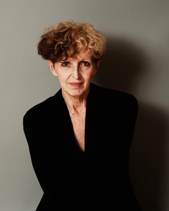 A woman with two toned curly hair sits on a gray background with a black blazer.