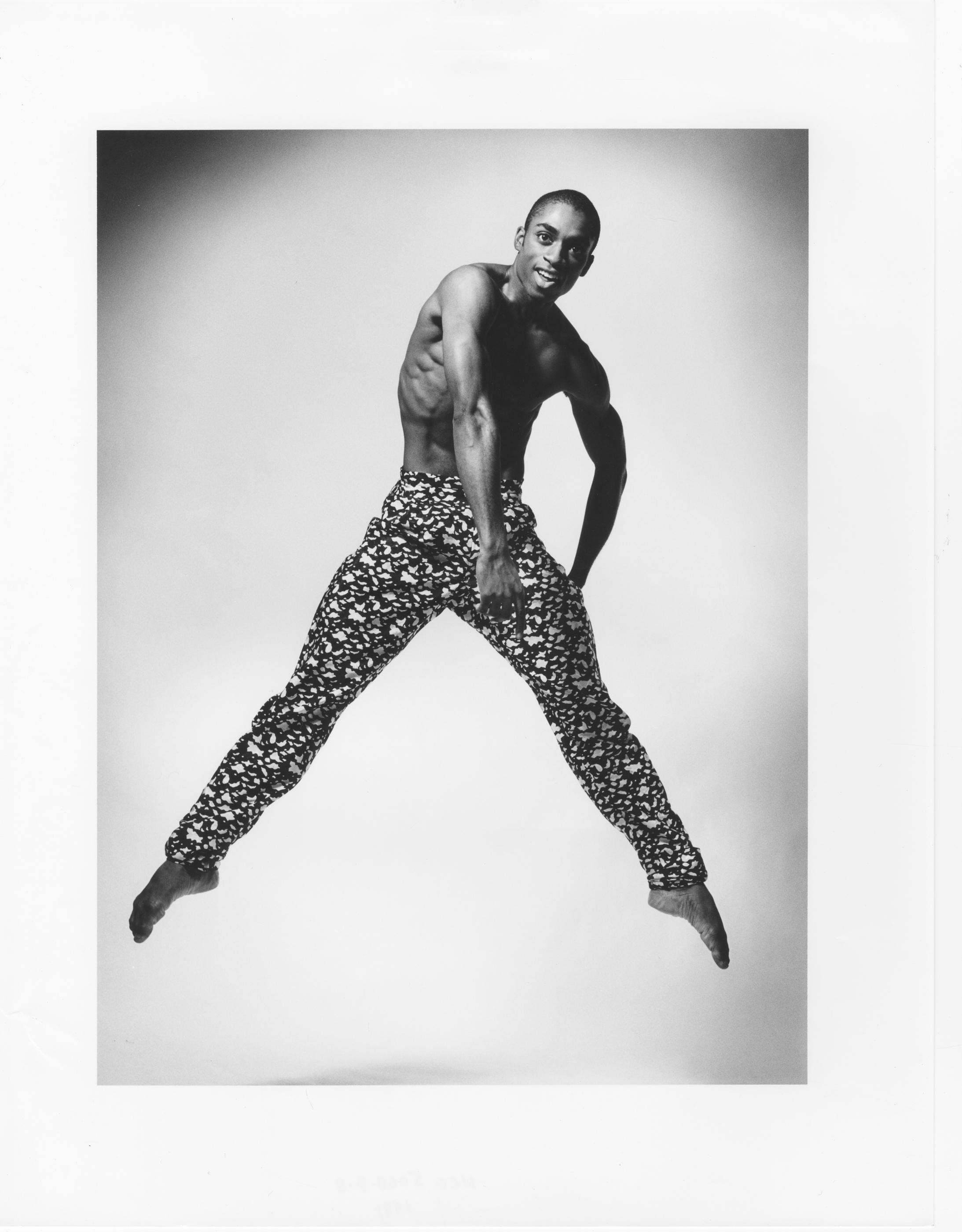 A black and white photo of dancer Desmond Richardson in mid-air. He is shirtless, wearing patterned pants with a black and white design. He has one arm bent at his side and the other extended downward, with legs apart. The background is plain and light-colored. The photo is labeled REPERTORY Desmond Richardson in Escargot. Photo by Jack Mitchell.