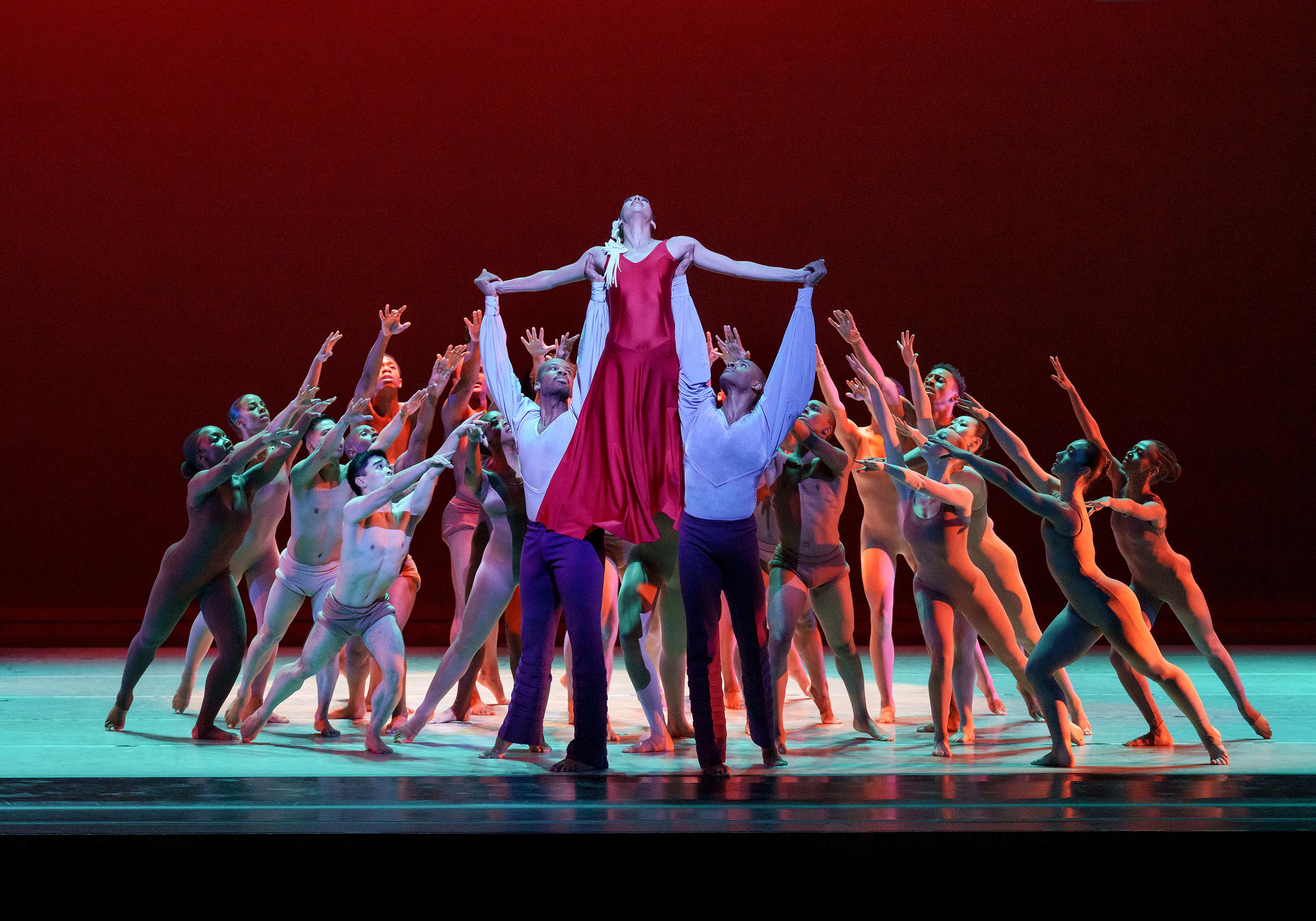 A group of dancers are huddled on stage forming a circle with their arms raised towards two men holding up one woman in a cross-like position. The woman is in red and the men are in white button downs and black pants.