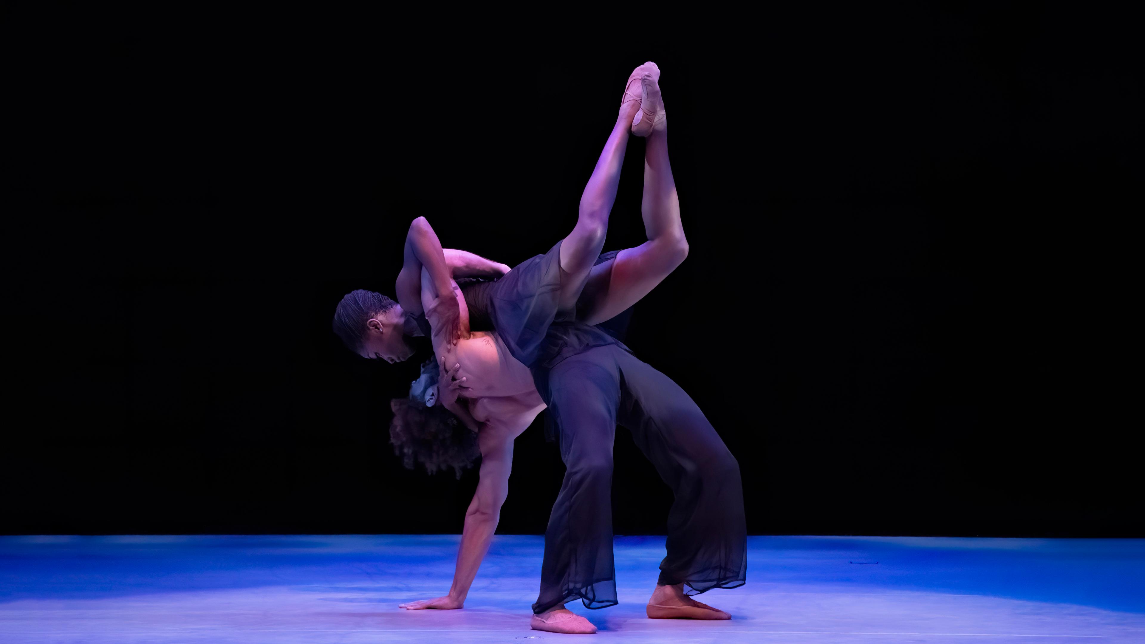 A man in a backbend with one hand on the ground and a women rests on his chest with her legs bent up in the air.