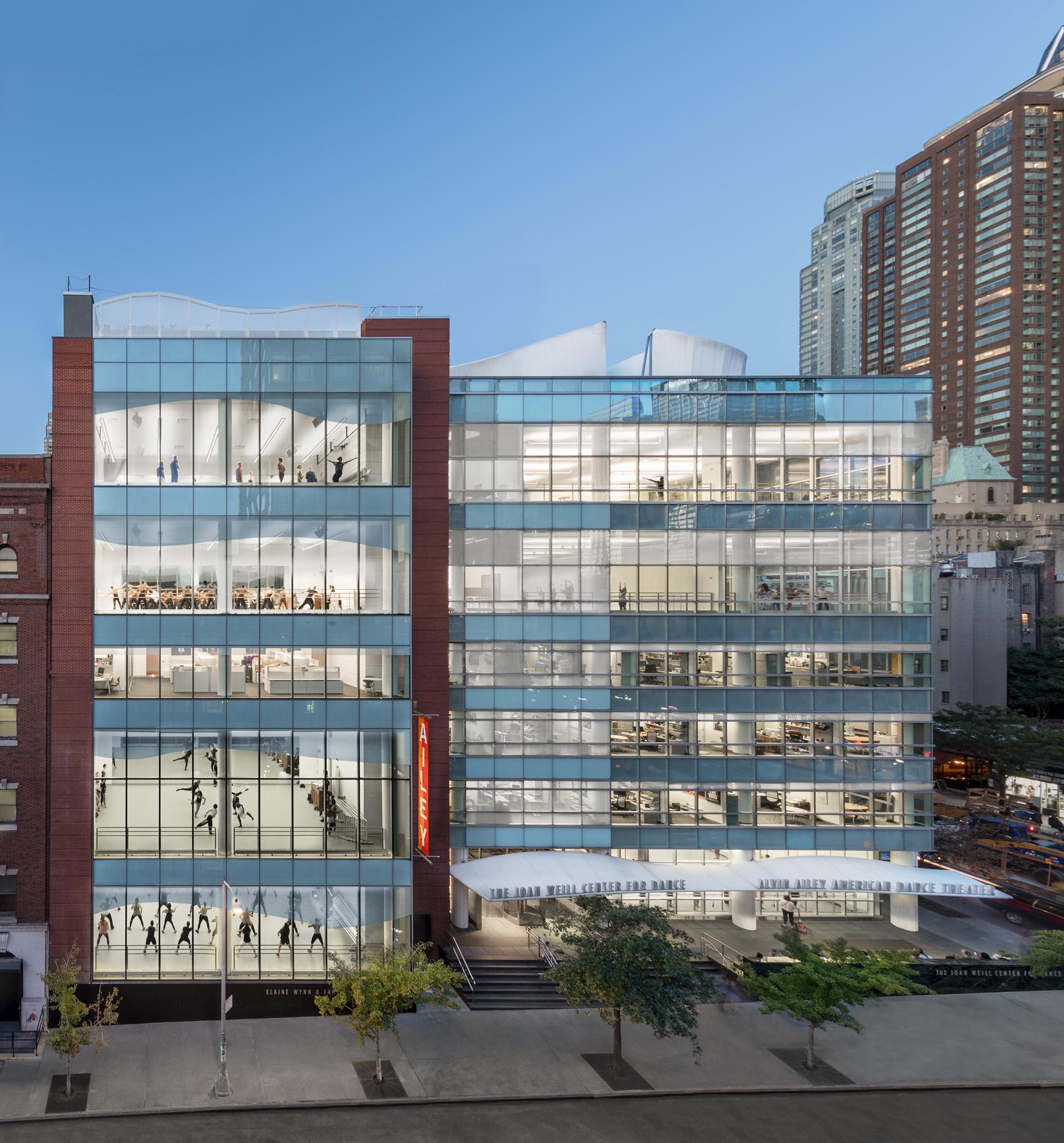 Exterior view of The Joan Weill Center for Dance with the Elaine Wynn & Family Education Wing. The modern building has large glass windows, showing multiple floors with people practicing dance inside. The building is surrounded by other tall buildings in an urban setting.