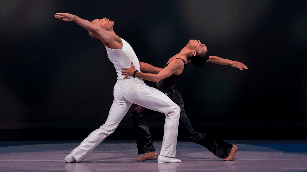 A male and female dancer hold each other on stage with arms in the air. She is in all black and he's in all white