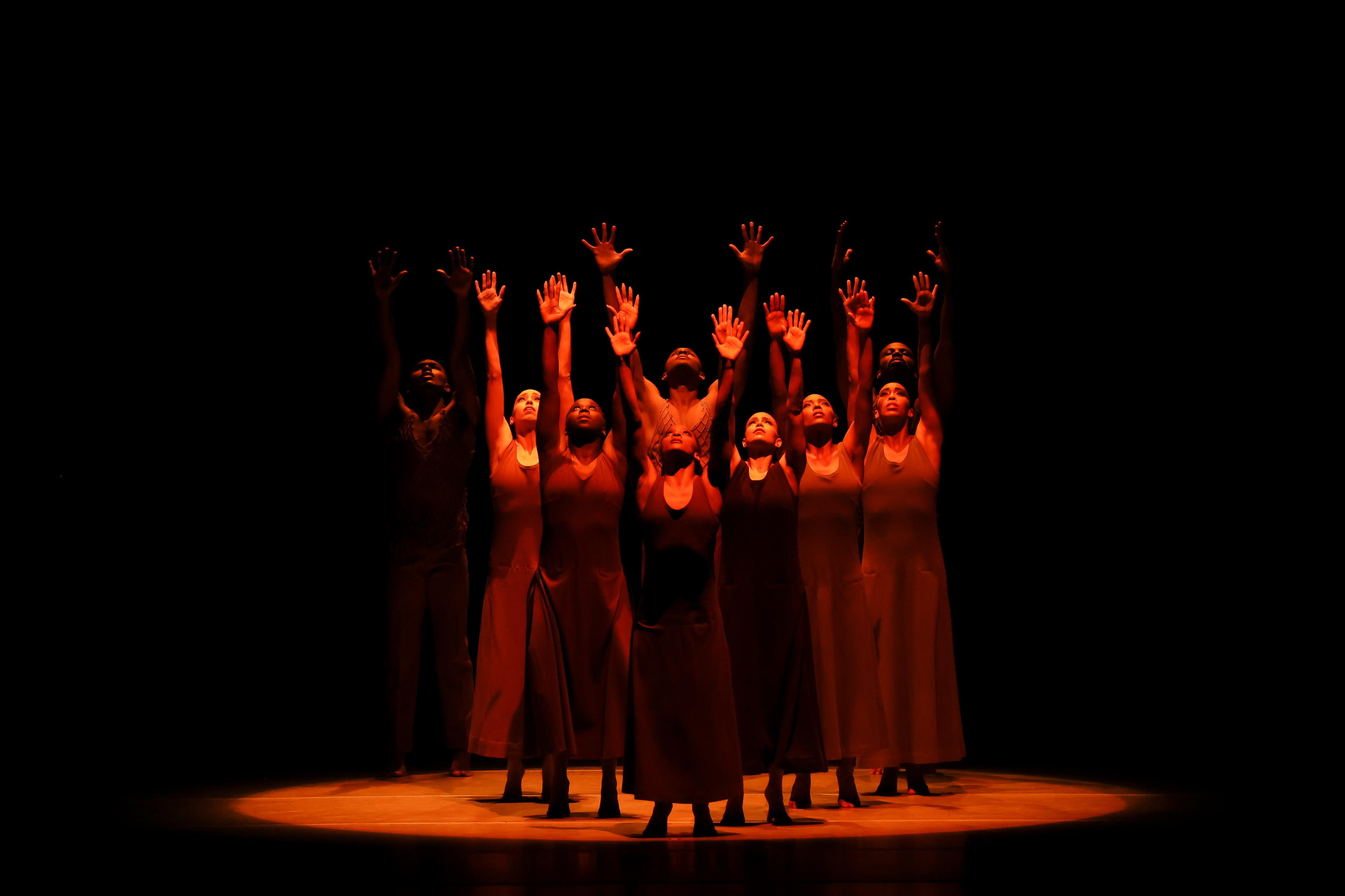 A group of dancers in flesh tone dresses look up and reach their hands to the sky 