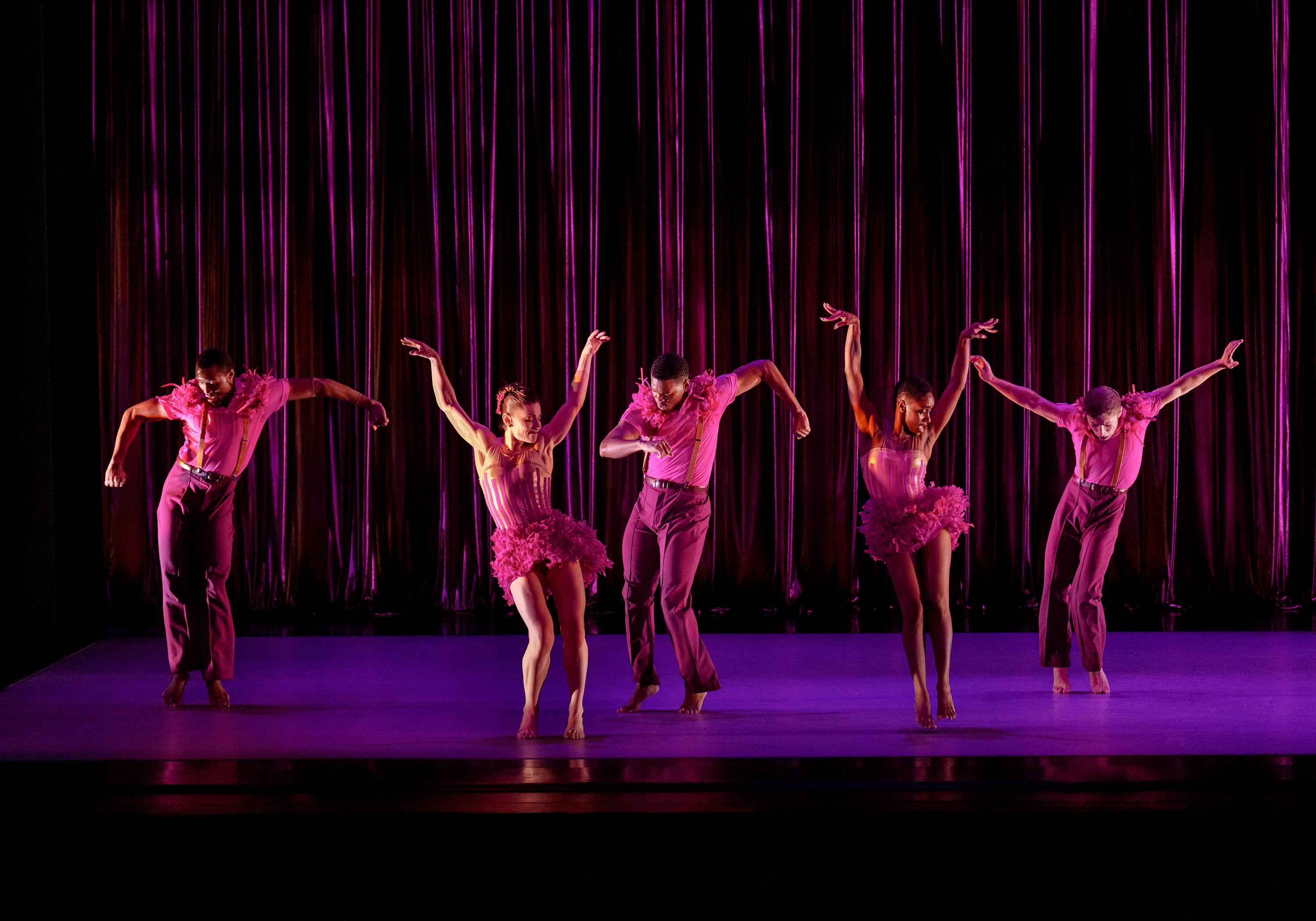 A group of dancers dressed in pink dresses and pink suites pose on stage