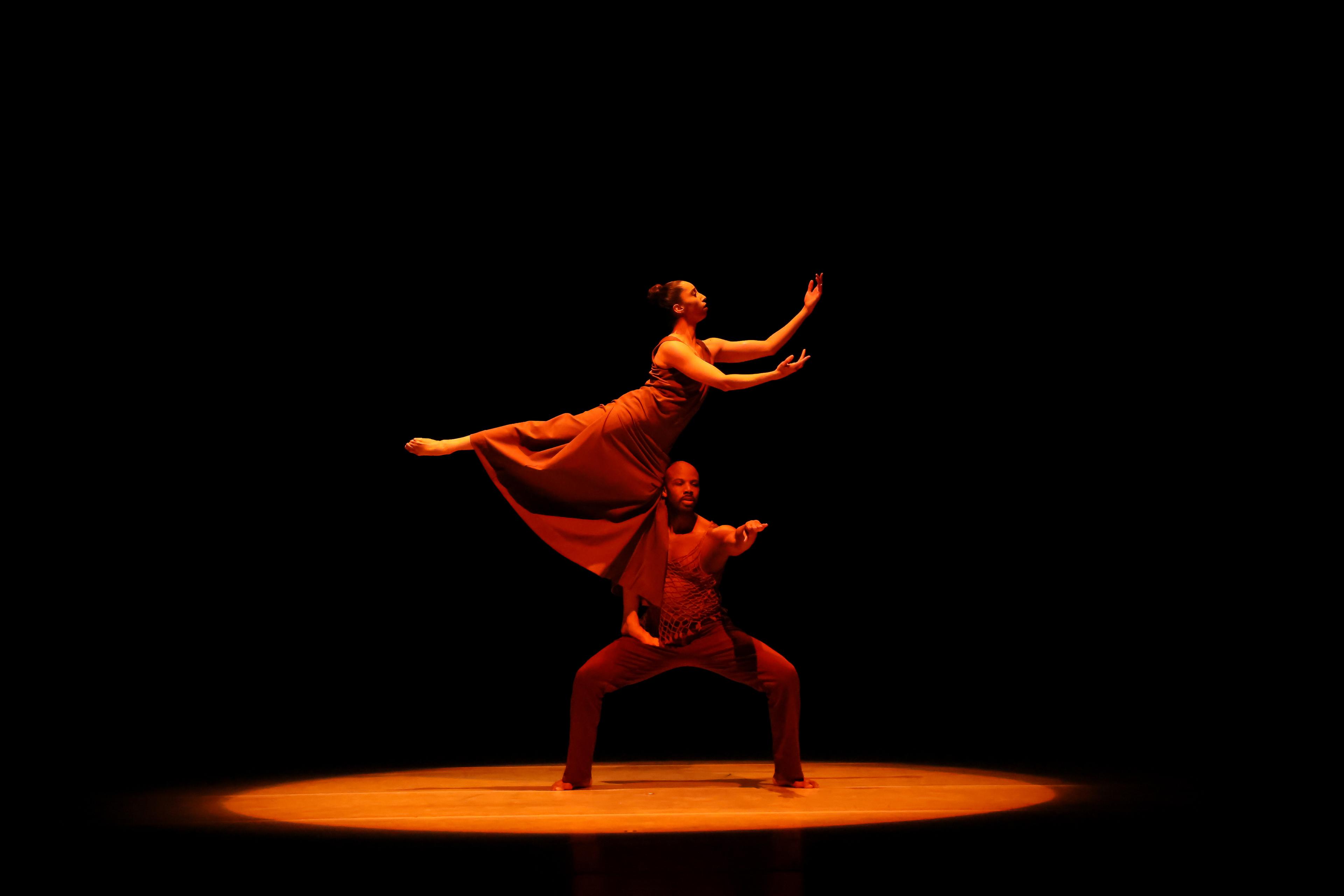 2 dancers in brown costumes, 1 squatting with the other standing on their leg in arabesque