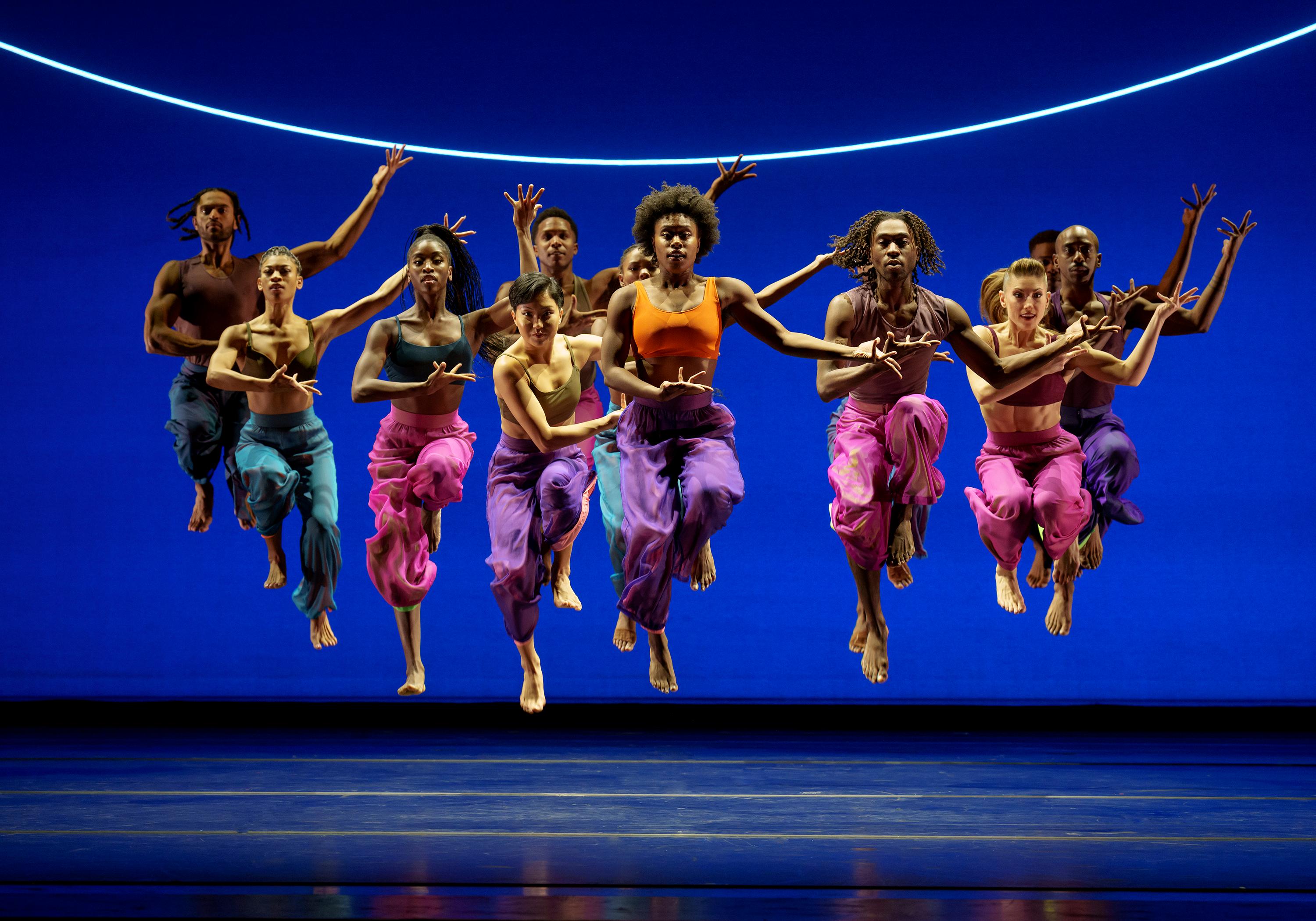 A group of dancers from the Alvin Ailey American Dance Theater performing "Are You in Your Feelings" by Kyle Abraham. They are captured in mid-jump, wearing colorful flowing pants and crop tops, set against a bright blue background with a curved light above them.