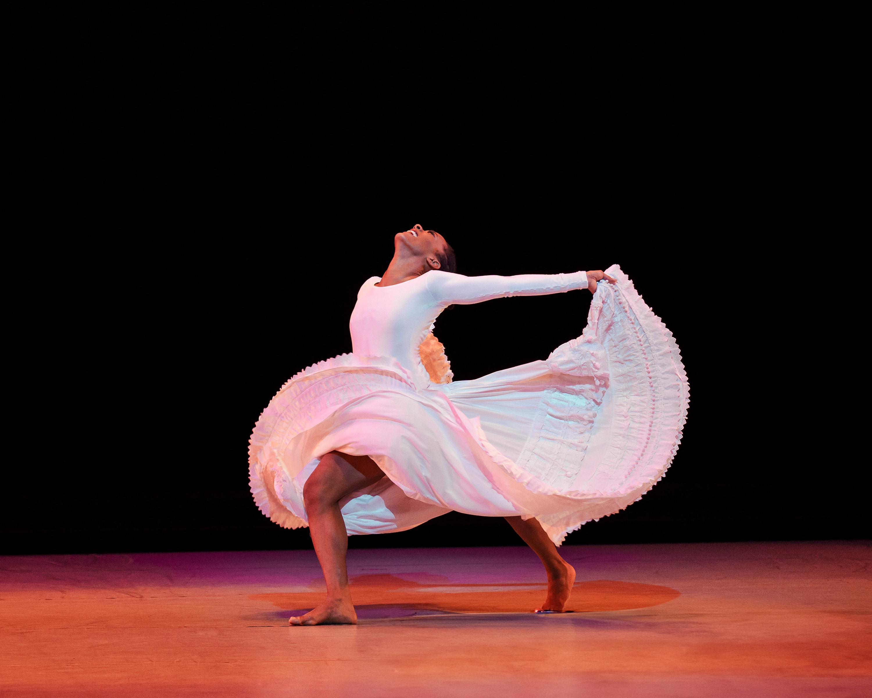 A dancer in a white long sleeve dress throwing her arms back while holding the dress