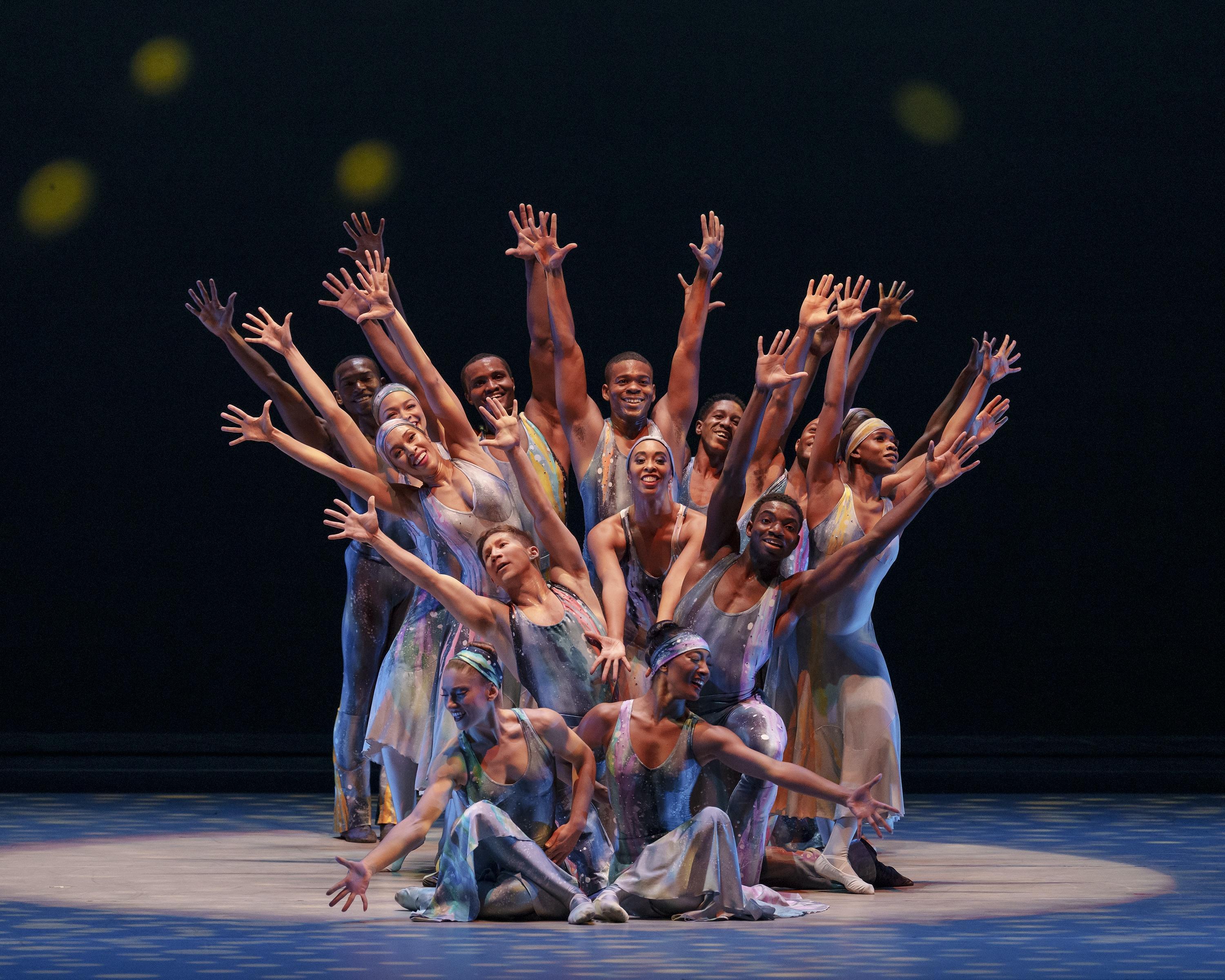 A group of dancers in colorful costumes pose with arms extended upward in a dynamic formation on stage. The background is dark with subtle yellow light spots, enhancing the vibrant energy of the performance.