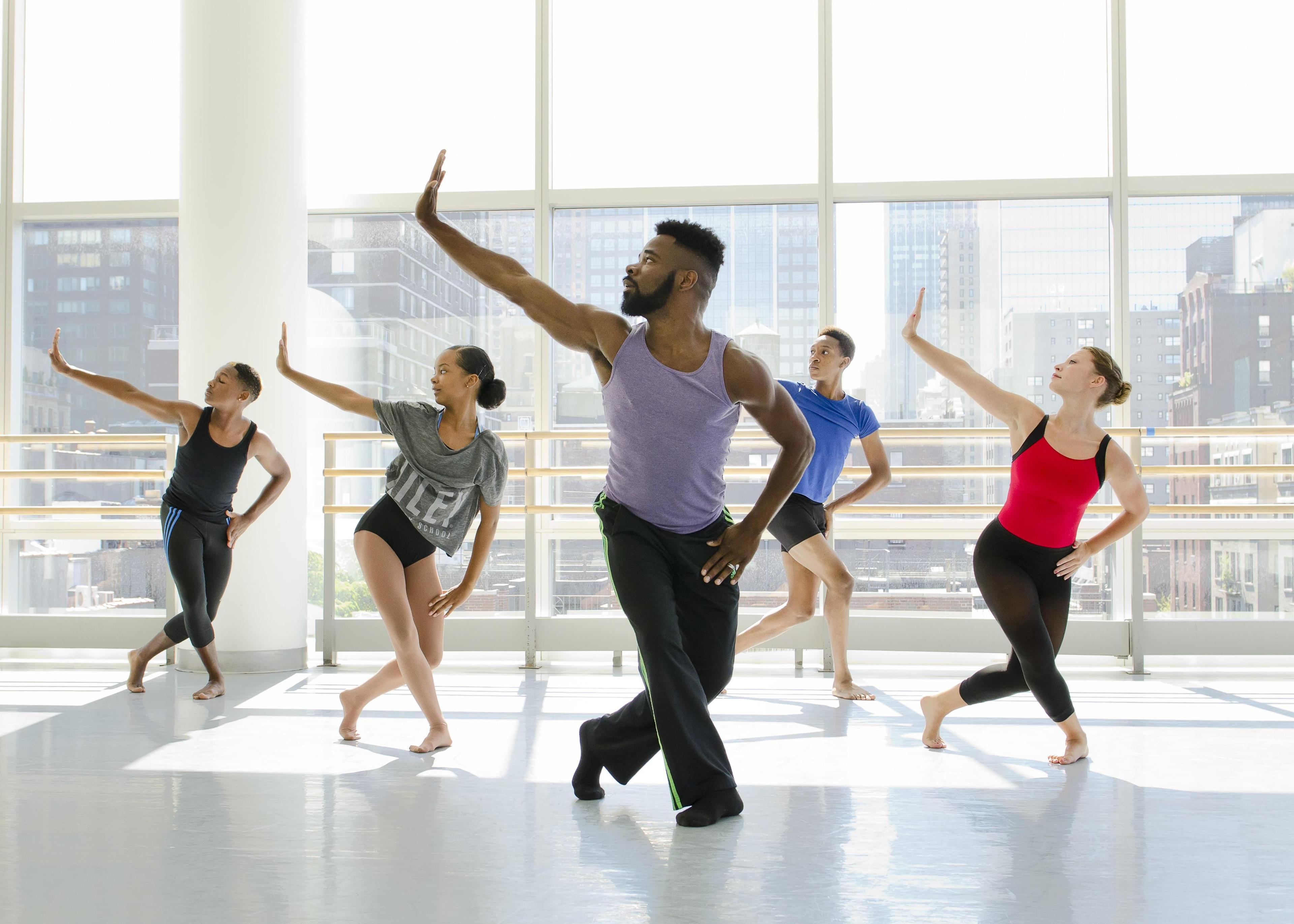 a dance class at The Ailey Extension. The dancers are mid-movement, each striking a pose that emphasizes fluidity and grace. They are positioned in a spacious, light-filled studio with large windows that offer a view of the cityscape. The dancers are diverse, dressed in comfortable dance attire, and exhibit strong, expressive movements. 