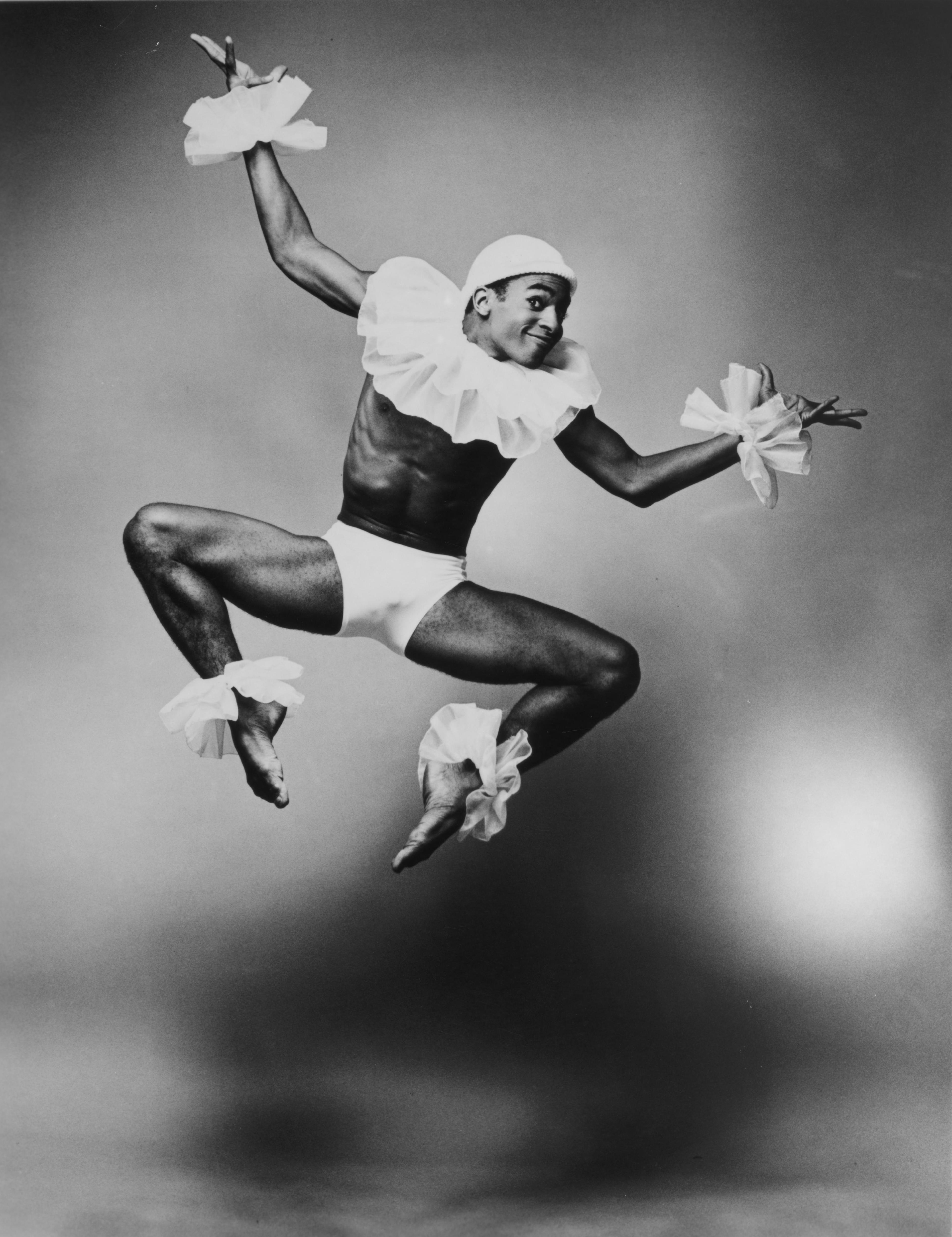 A black and white photo of a male dancer in mid-jump. He is wearing a white cap, a ruffled collar, wrist cuffs, ankle cuffs, and briefs. The background is light.