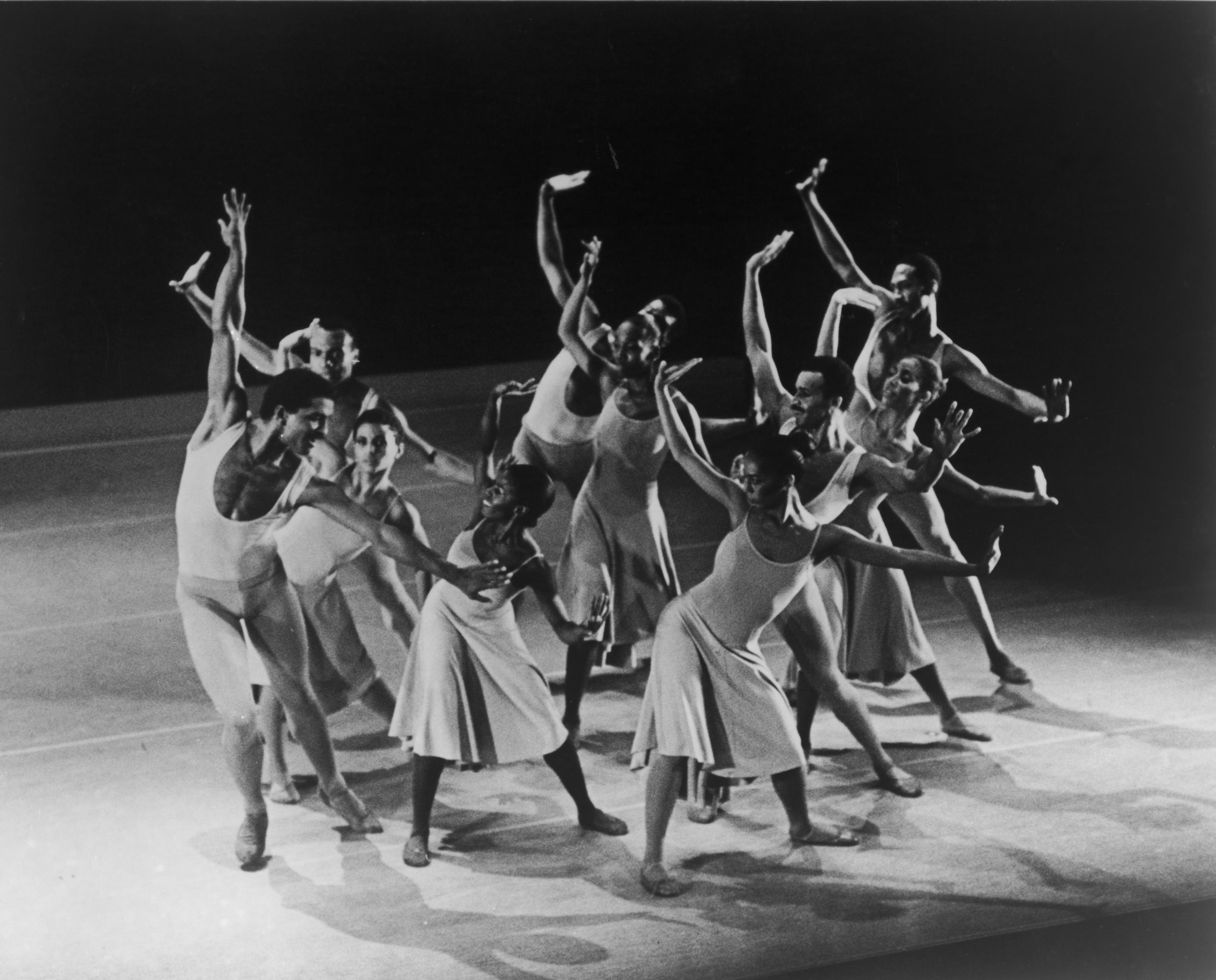 A black and white photo of a group of dancers in mid-performance, wearing light-colored costumes. They are striking dynamic poses with arms raised and legs extended. The background is dark, highlighting the dancers. The photo is labeled REPERTORY Concerto in F. Photo by Jack L. Vartoogian.