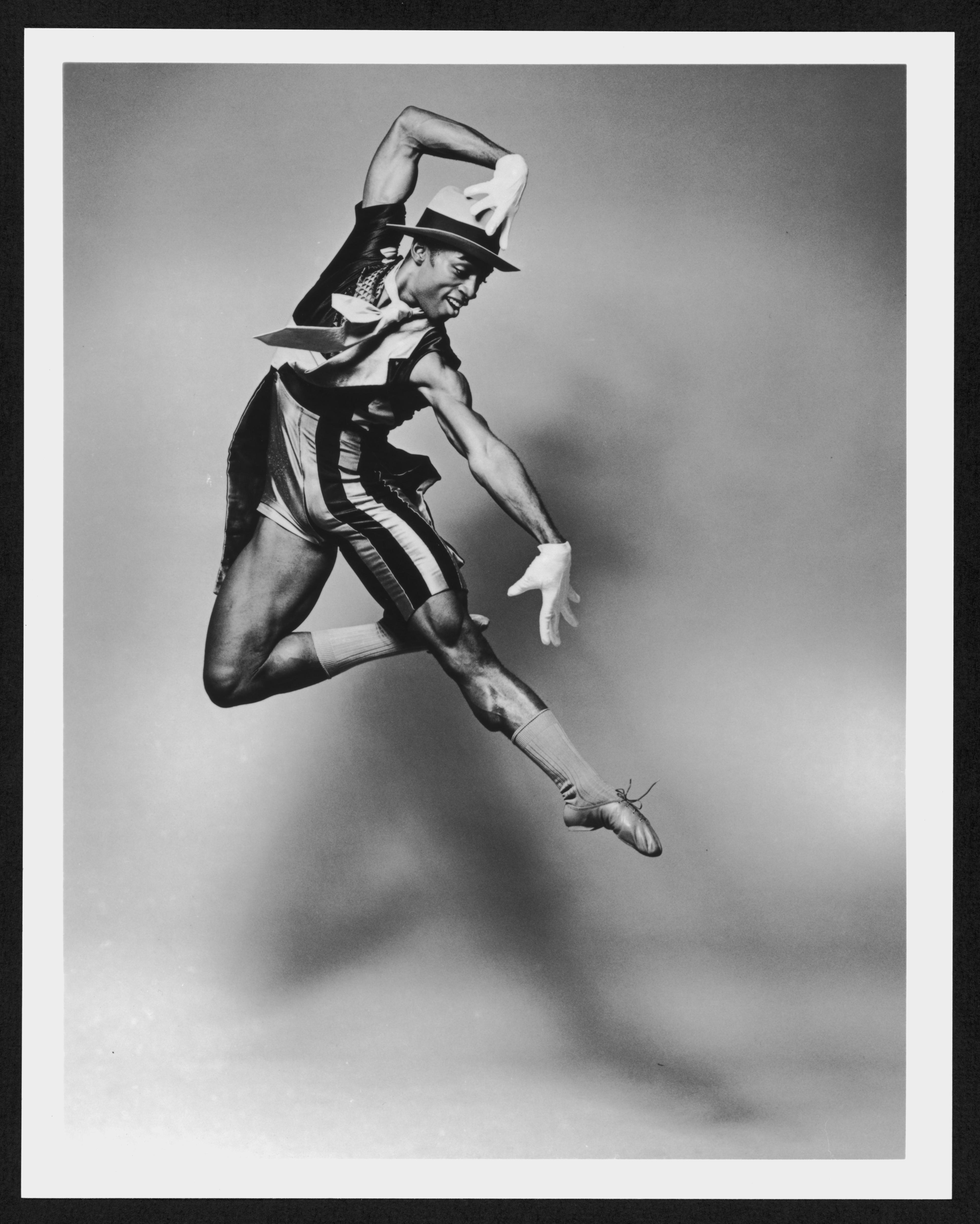 A black and white photo of dancer Desmond Richardson in mid-air. He is wearing a costume that includes a hat, gloves, a striped vest, and shorts. One arm is bent above his head, and the other is extended downward, with one leg bent and the other extended back. The background is plain and light-colored. The photo is labeled REPERTORY Desmond Richardson in Fontessa and Friends. 