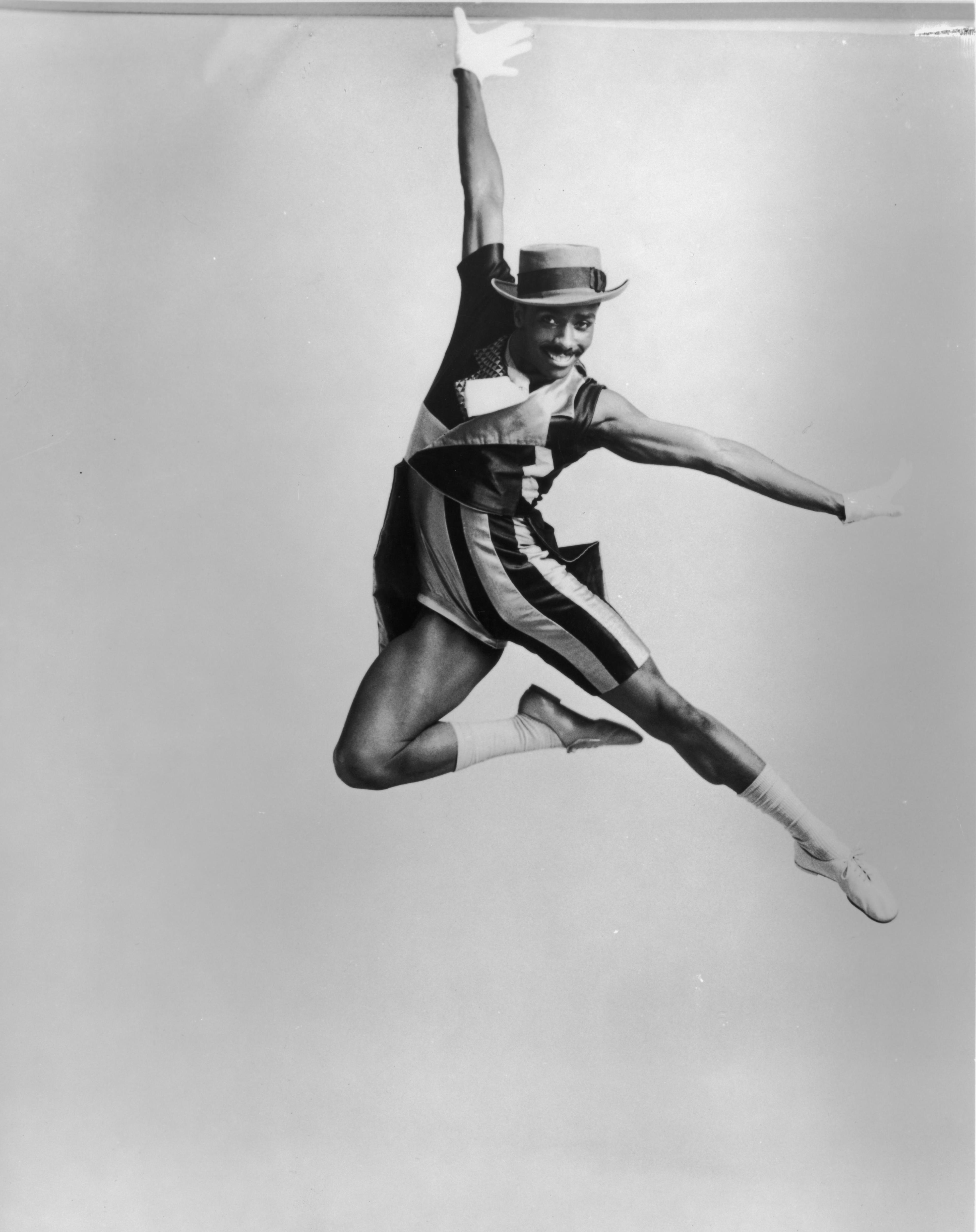 Black and white photo of Ralph Glenmore performing in Fontessa and Friends. Ralph is captured mid-jump with one arm raised and the other extended to the side, wearing a striped costume and a hat, and smiling.