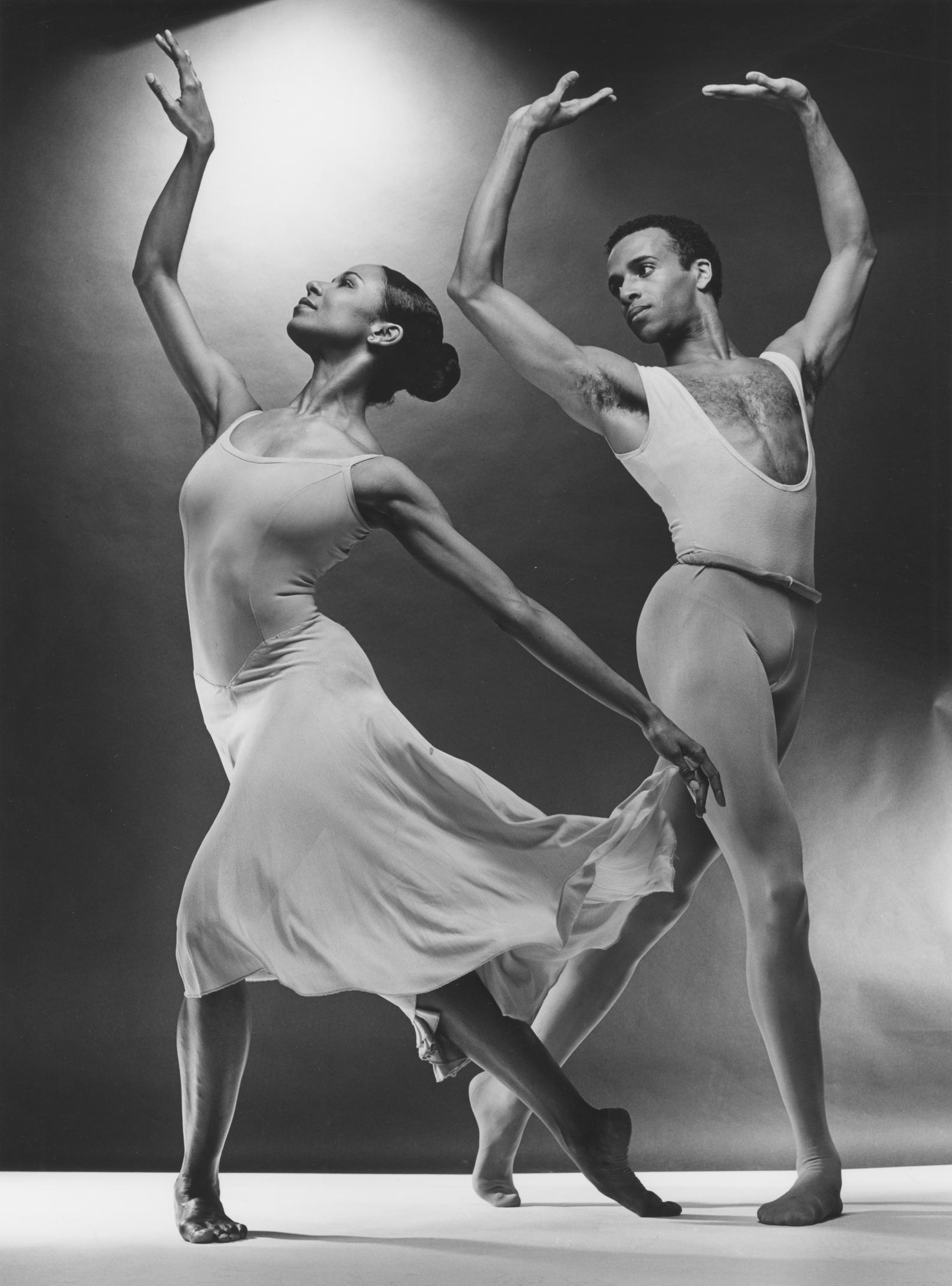 Black and white photo of Sara Yarborough and Carl Bailey performing in Concerto in F. Sara wears a flowing dress, and Carl is in a sleeveless leotard. Both dancers are captured in an elegant pose with arms gracefully raised.