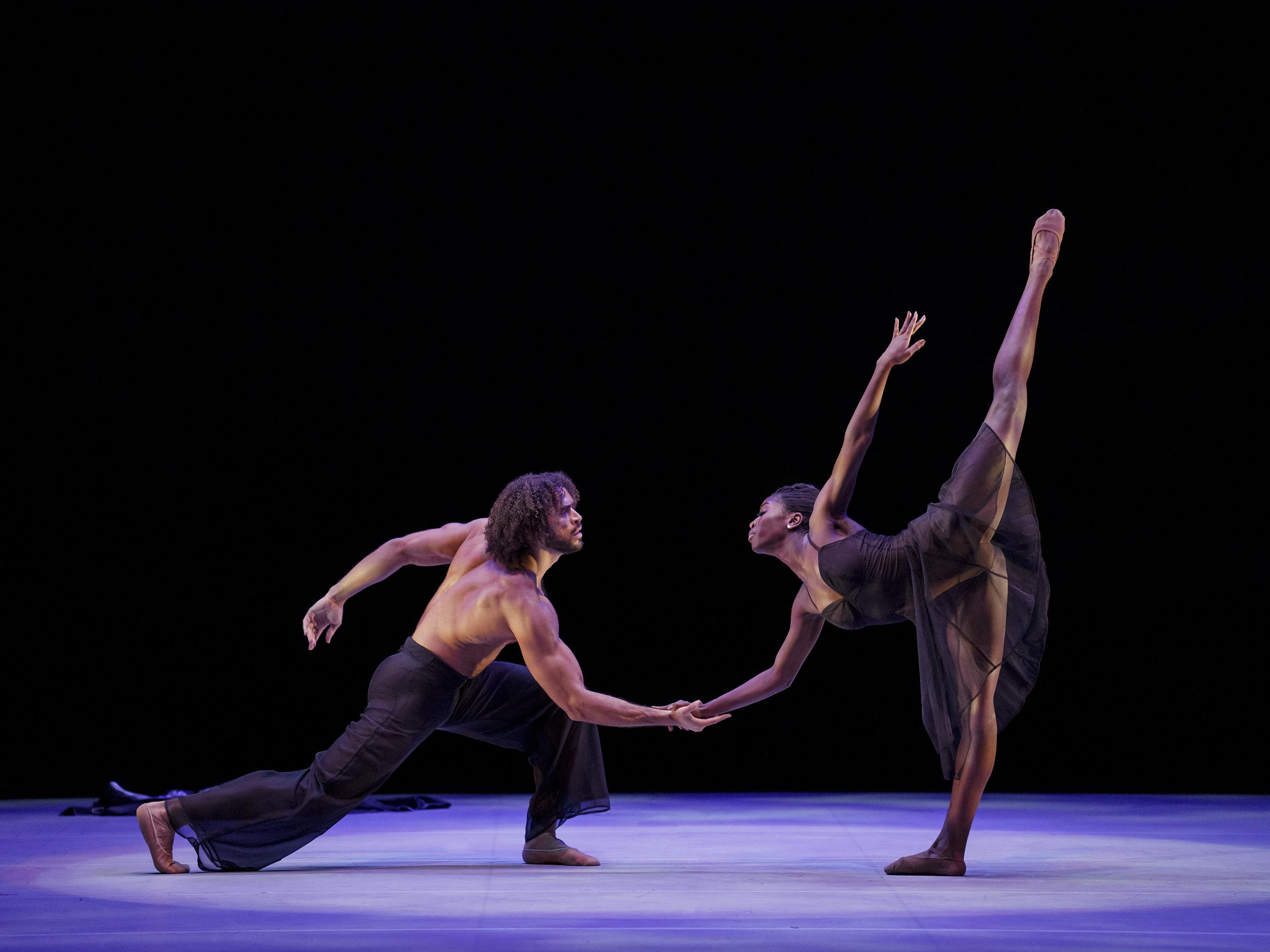 A shirtless man lunges forward holding the hand of a woman who is also bent forward standing in arabesque.