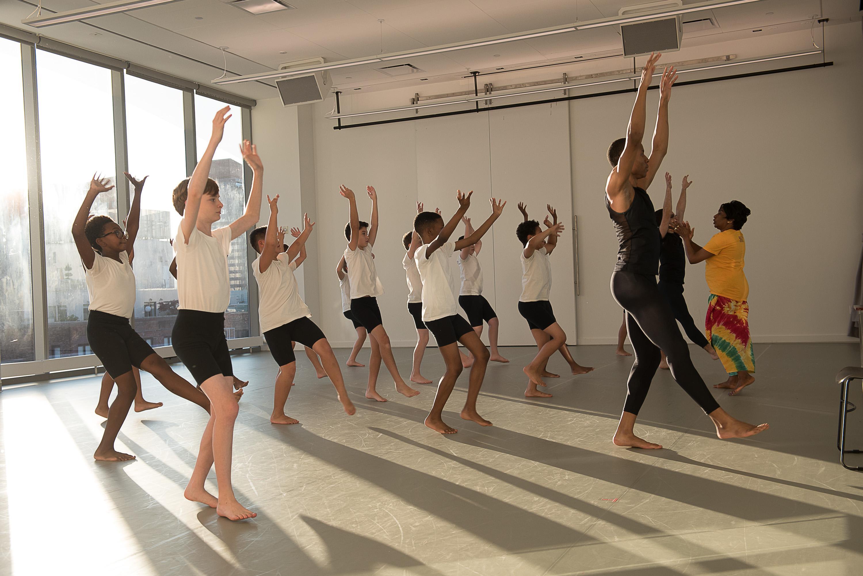  A group of boys in a dance studio wearing white shirts and black shorts, practicing a West African dance class. They are guided by two instructors, one in black and the other in a yellow shirt and colorful skirt. The studio has large windows letting in natural light.