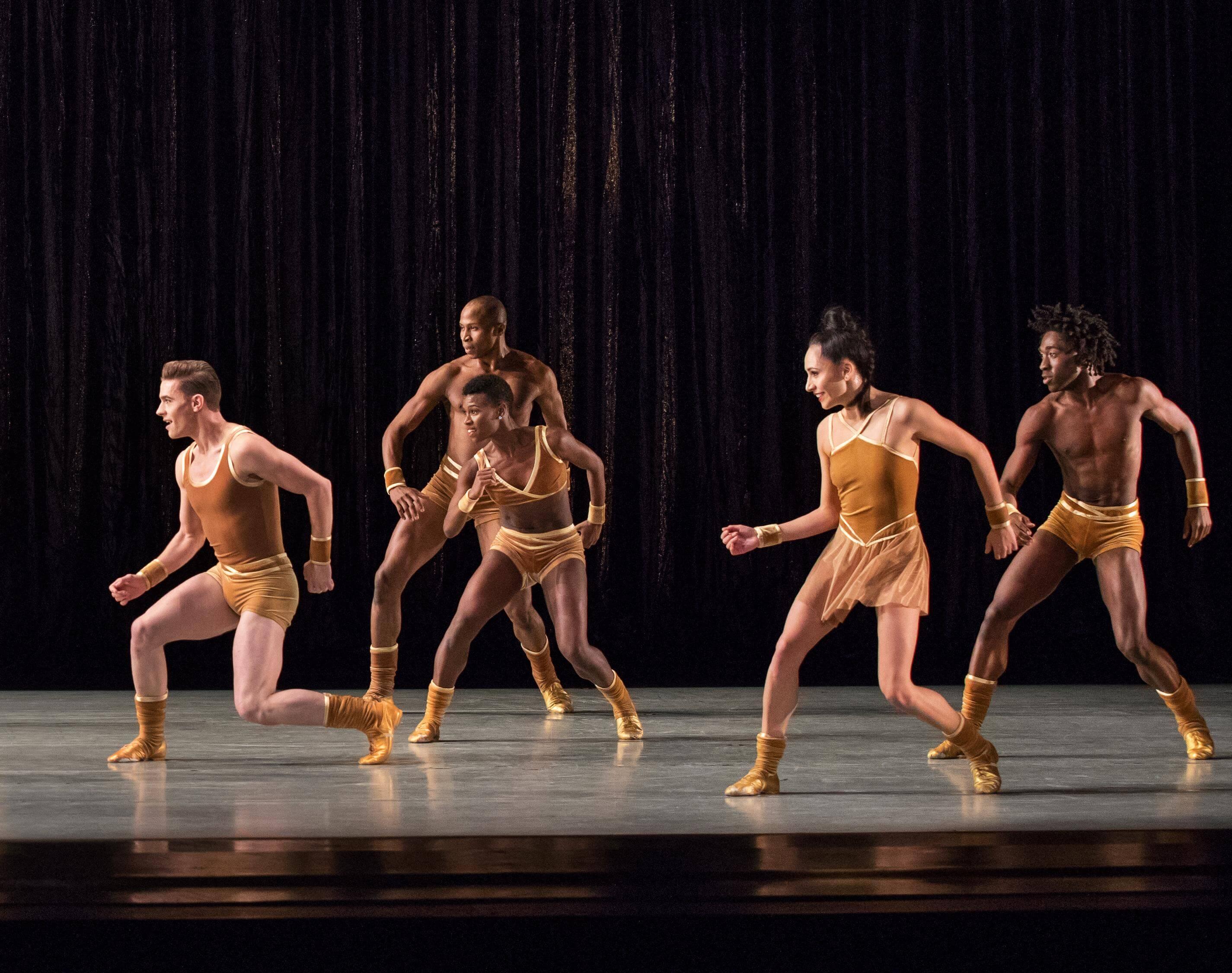 Five dancers in yellow costumes perform a synchronized dance on stage. They are positioned dynamically with bent knees and arms, emphasizing movement. The background is dark, focusing attention on the dancers.