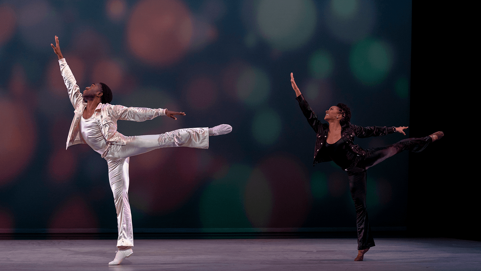 Two dancers, male in all white and a woman in black, are in arabesque on stage