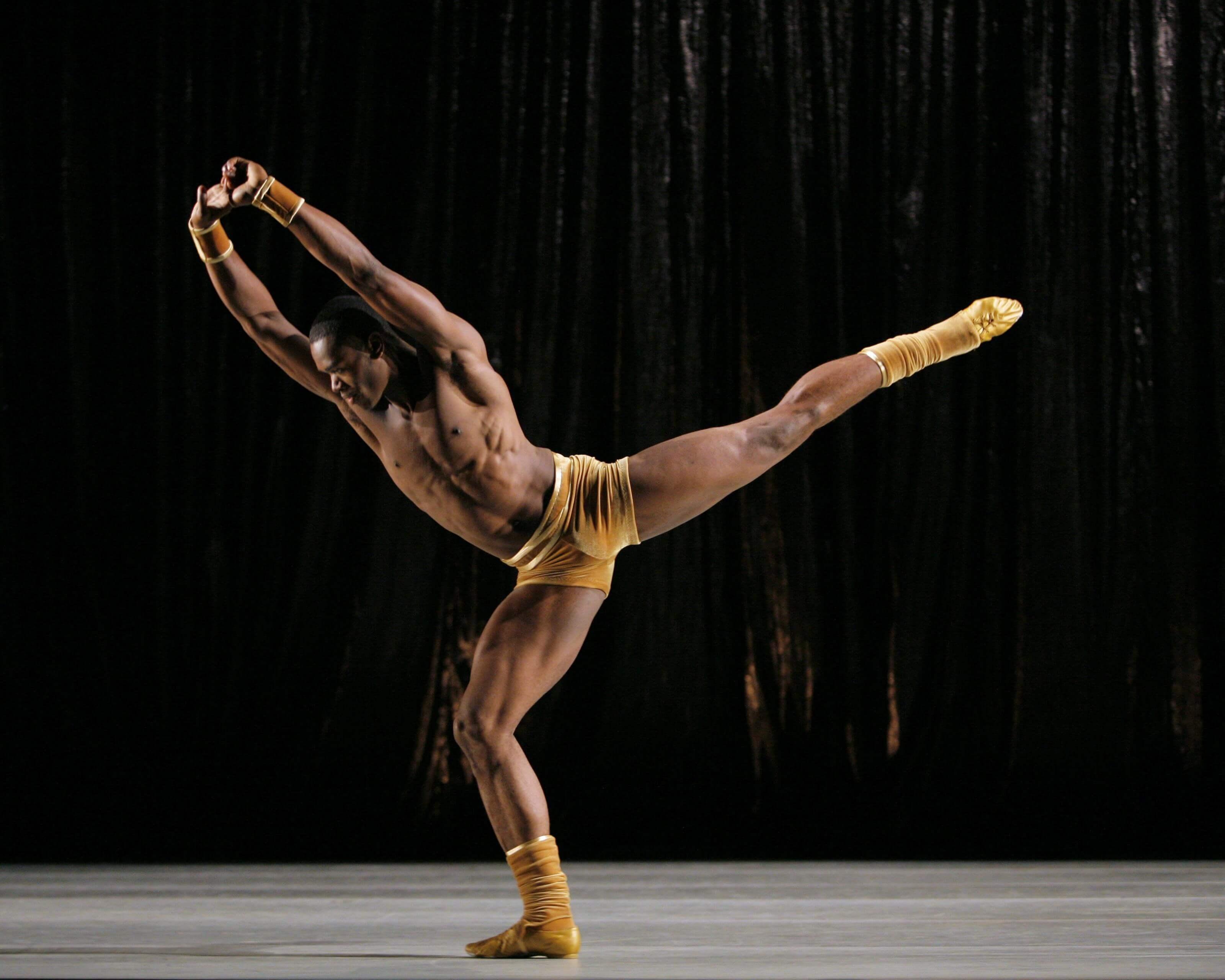  A dancer in yellow attire performs a high arabesque on stage. The background is dark, highlighting the dancer's muscular form and extended leg.