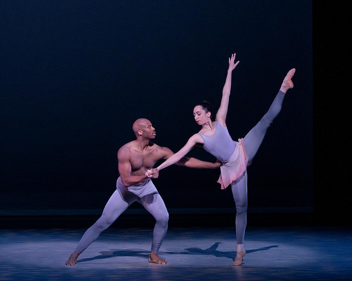 A color photo of Jermaine Terry and Sarah Daley-Perdomo performing in Alvin Ailey's The River. Terry, shirtless and wearing light-colored tights, supports Daley-Perdomo, who is wearing a pastel-colored leotard and skirt, as she extends one leg gracefully in an arabesque position, set against a dark background.