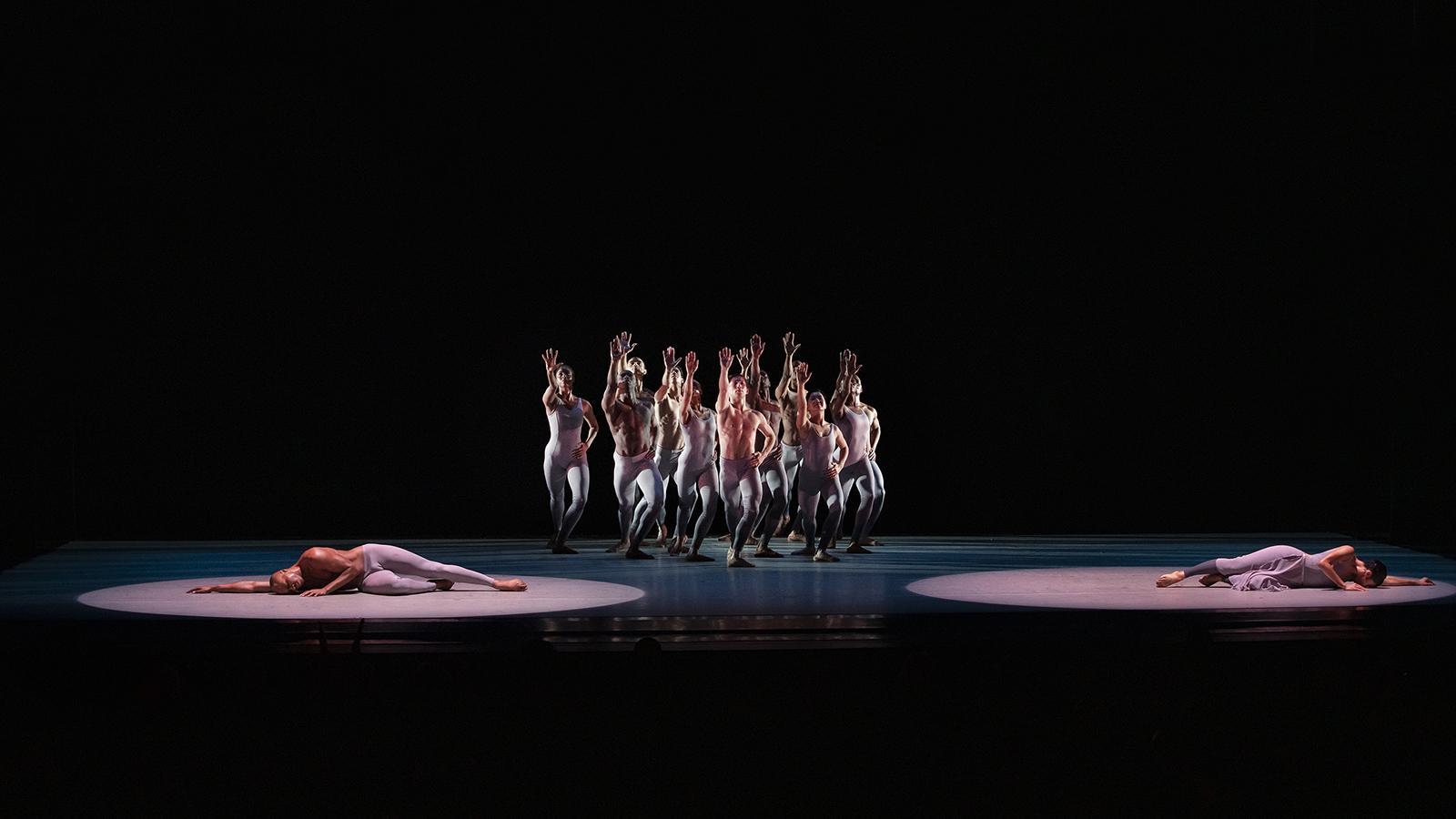 A color photo of the Alvin Ailey American Dance Theater performing Alvin Ailey's The River, captured by Paul Kolnik. The image features a group of dancers in the center, all wearing light-colored costumes and standing in a formation with their hands raised. Two dancers are lying on the stage, each under their own spotlight, on either side of the group. The background is dark, which highlights the dancers in the spotlight.