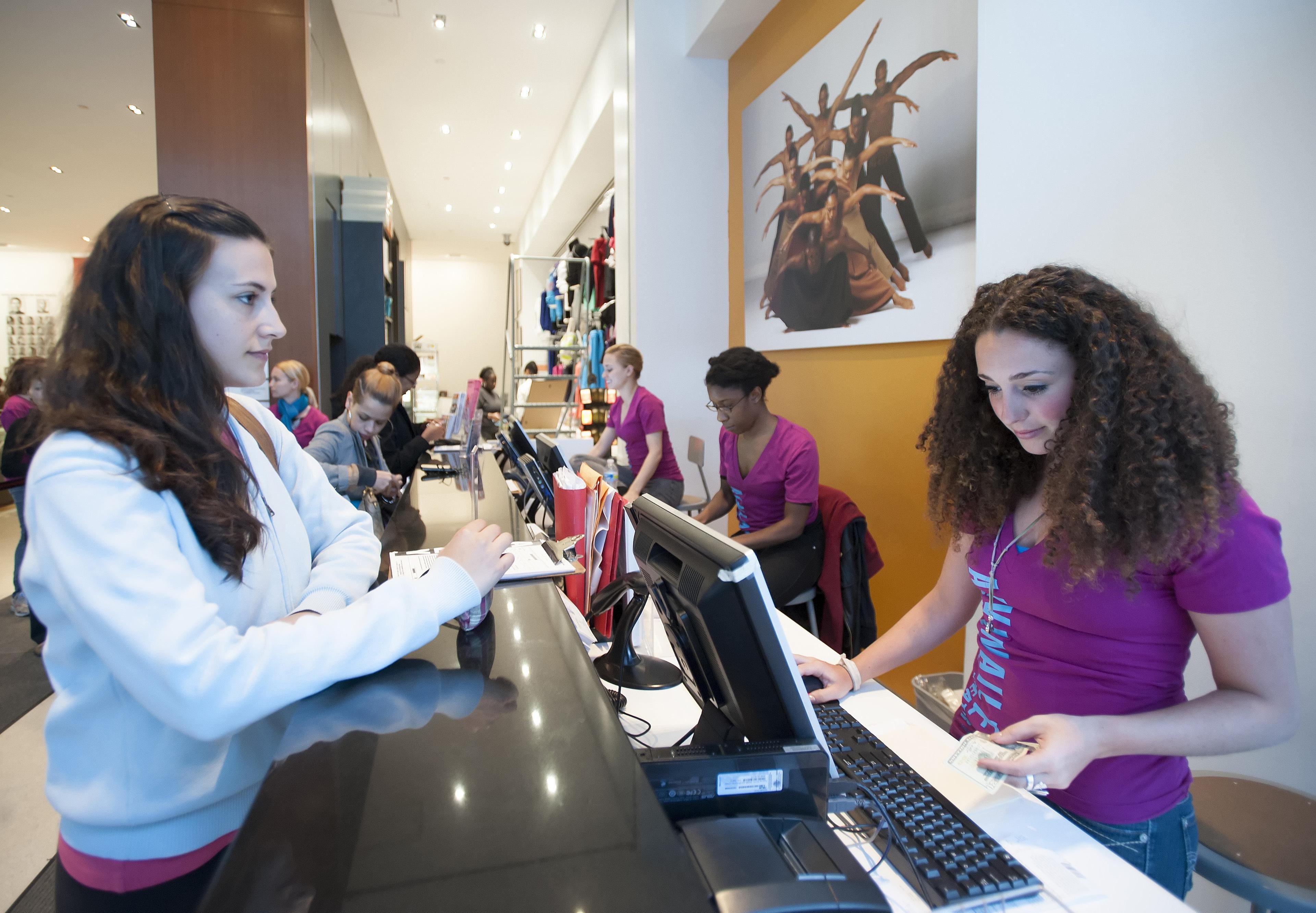 A woman checks in at the front desk of a studio. Women behind the desk are in purple shirts.