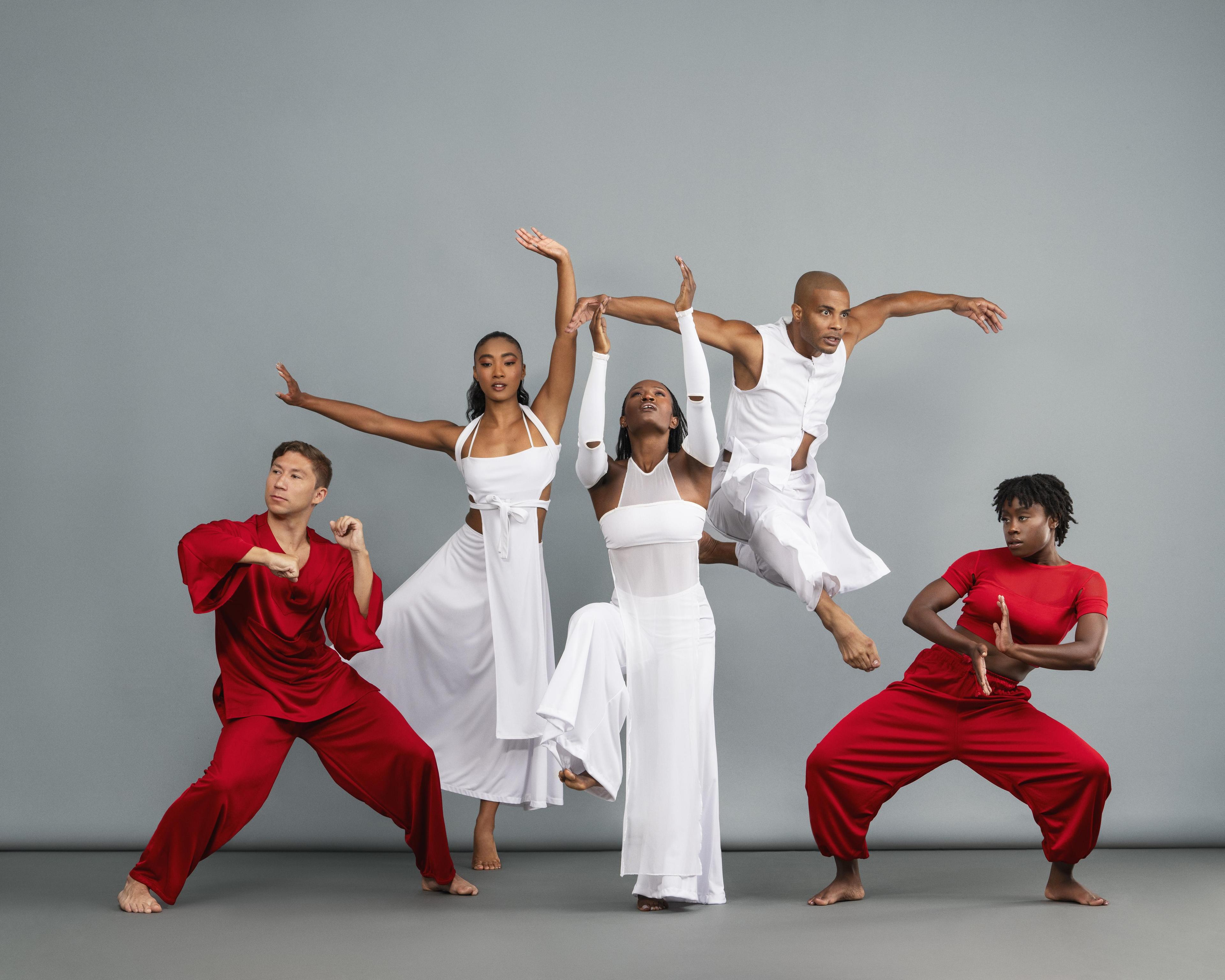 Five dancers. Three are in white and two are in red. They are standing in different poses, and one is jumping in the background. They are in a studio with grey walls.