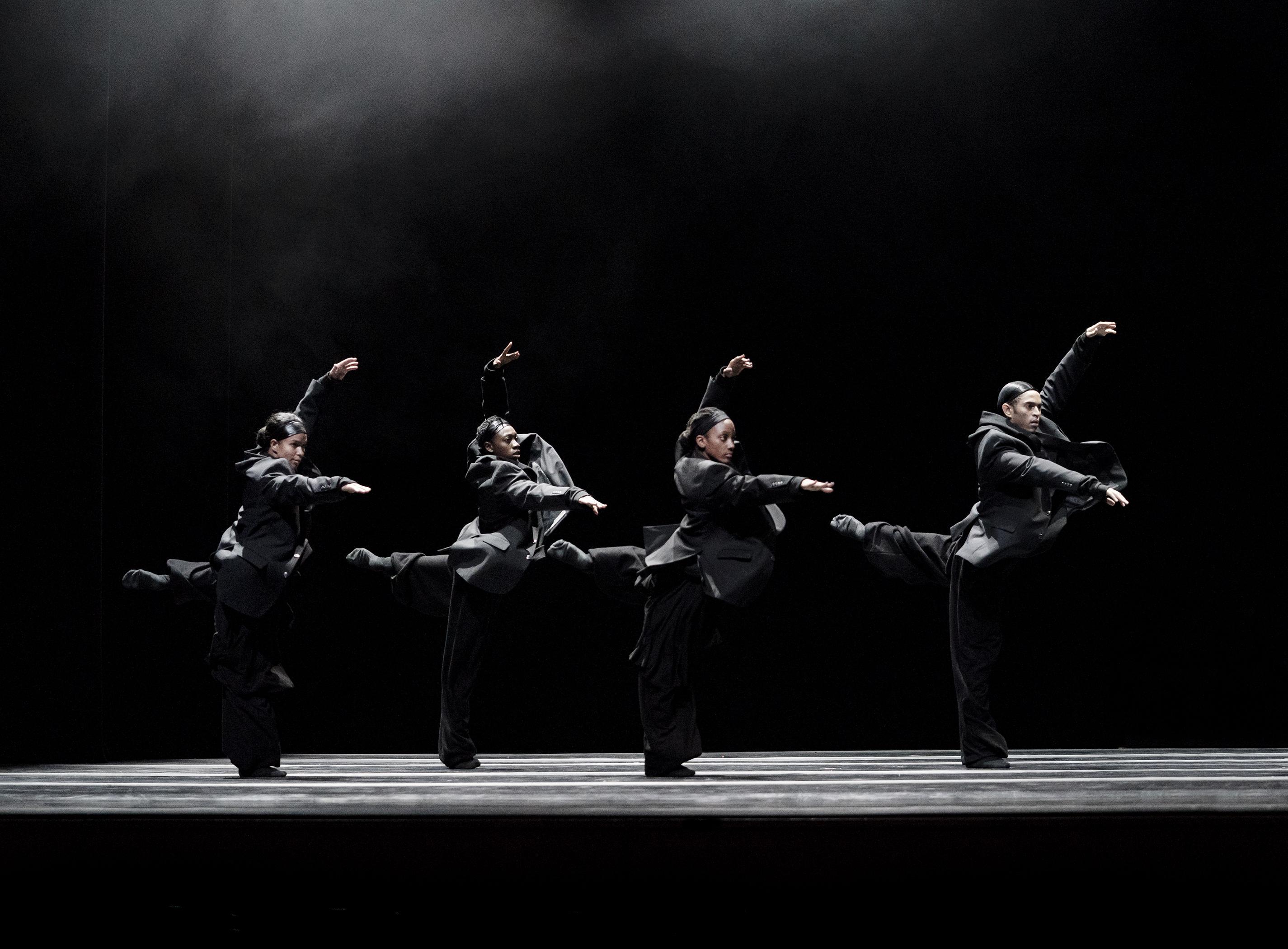 A group of four dancers performing the same pose with their leg extended backwards their arms above and in front of them