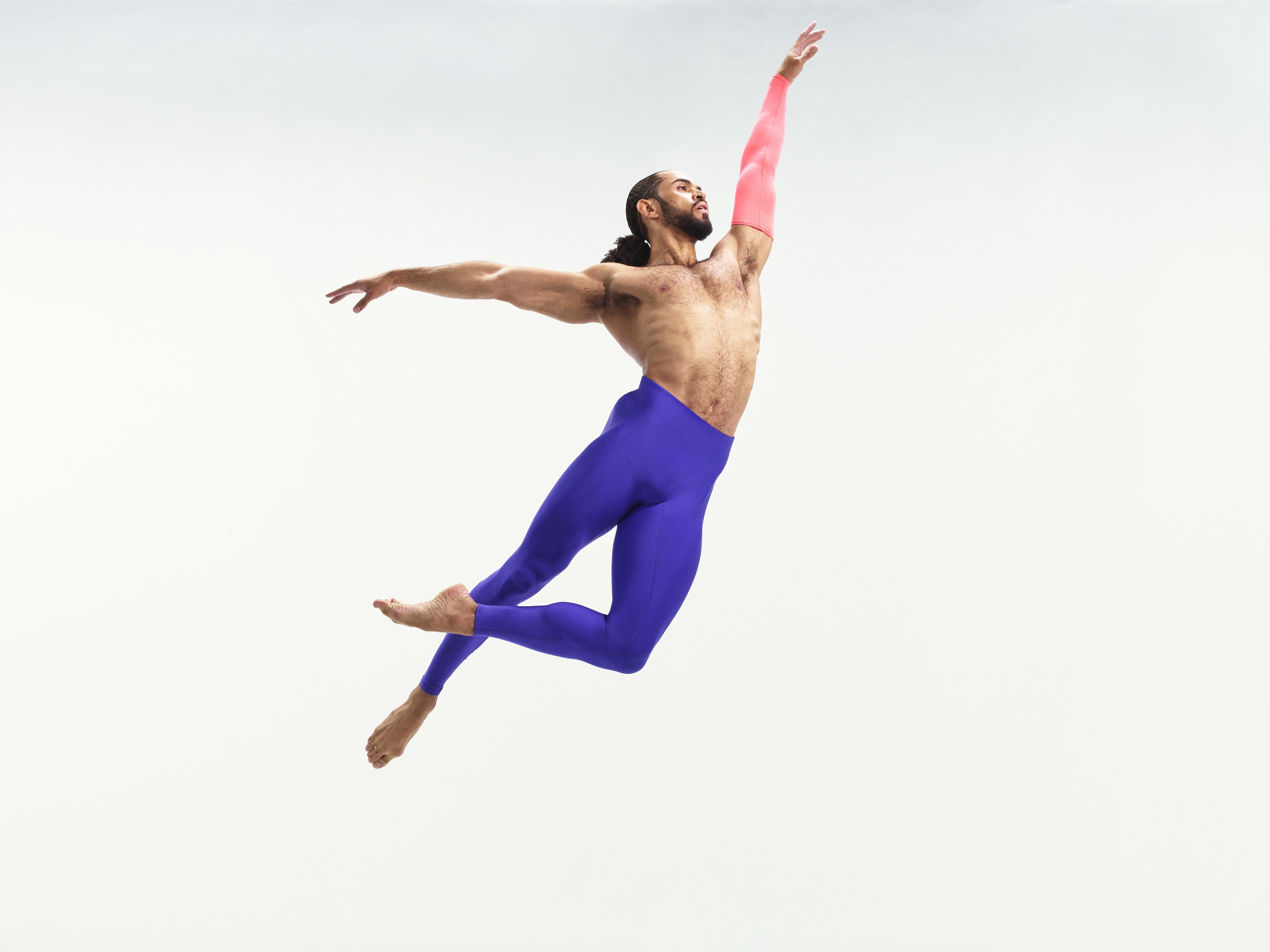 A shirtless male dancer in purple pants jumps up in the air