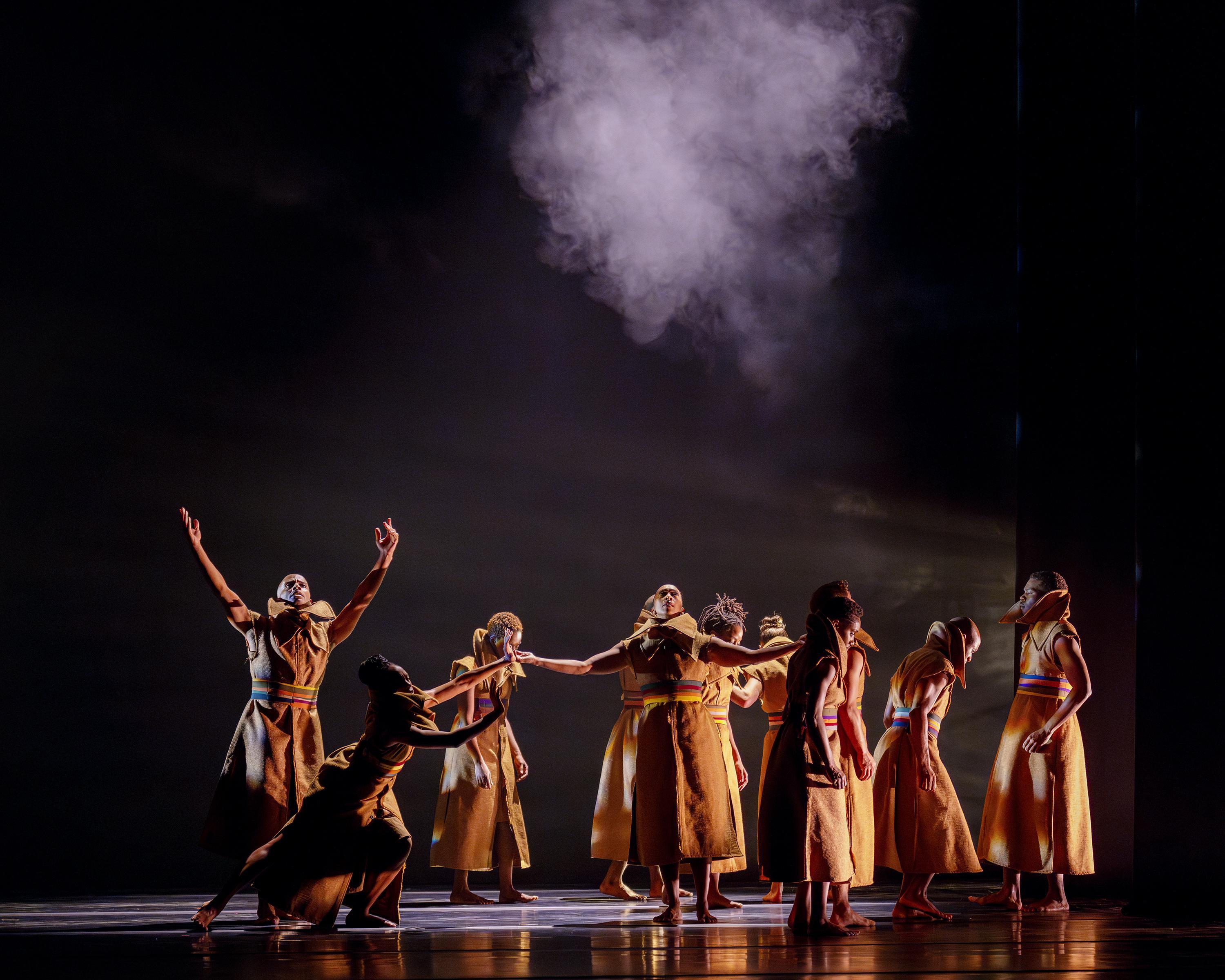 Dancers in dresses on a dark stage. There is a puff of smoke just above them.