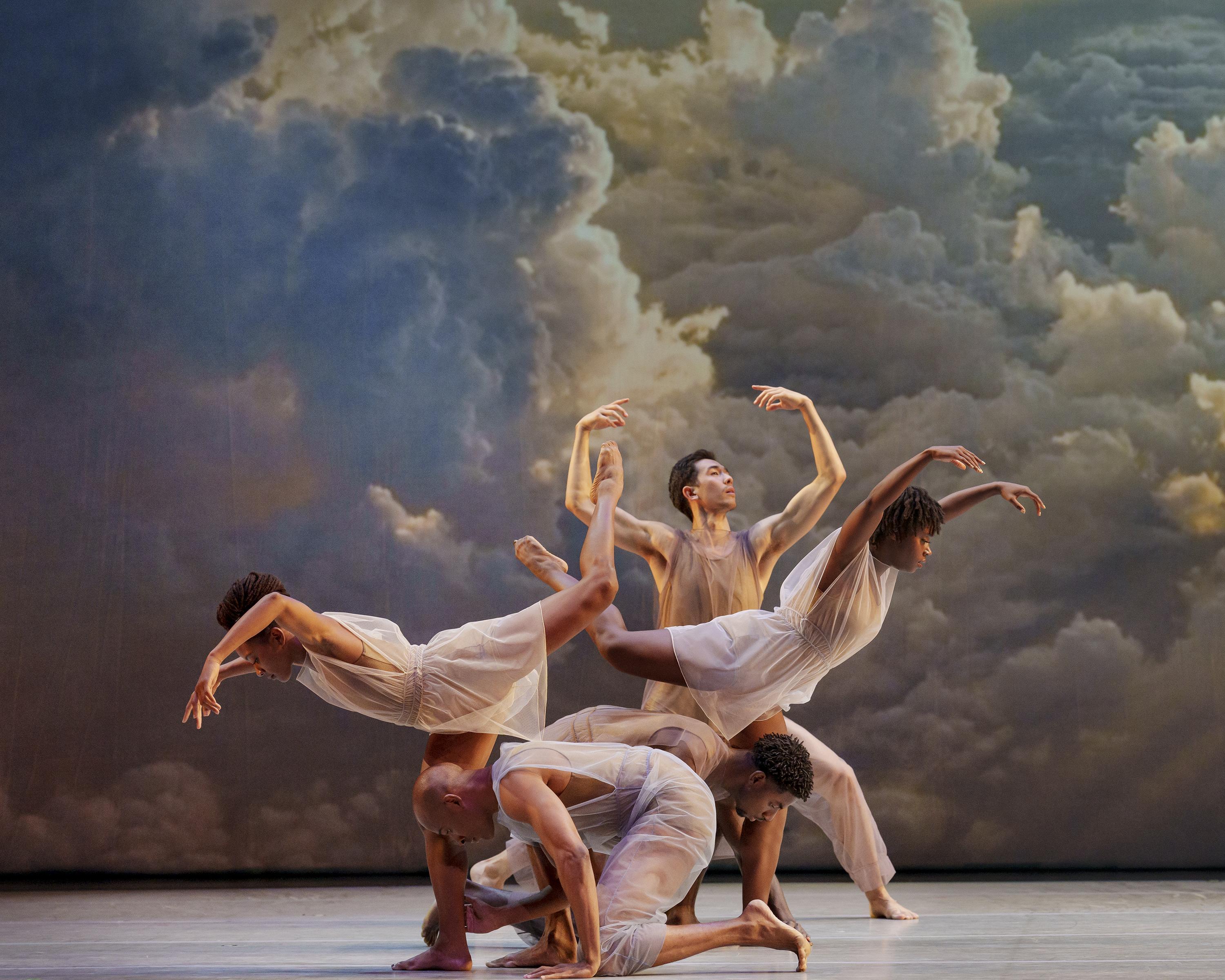 Dancers in white translucent costumes in a group with colorful clouds behind them