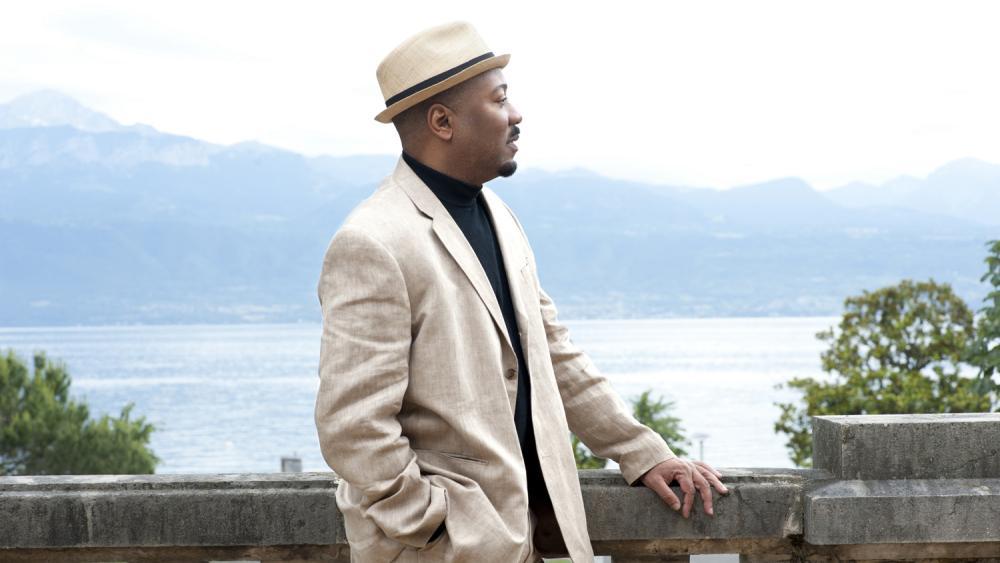 A black man stands with a lake behind him. He is in a white fedora and a white blazer. His arm rests on a railing.