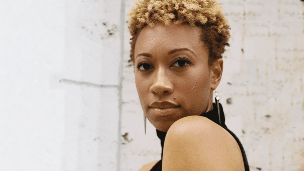 A black woman stands in front of a white brick wall. You can see her left shoulder but her face is looking at the camera straight on.