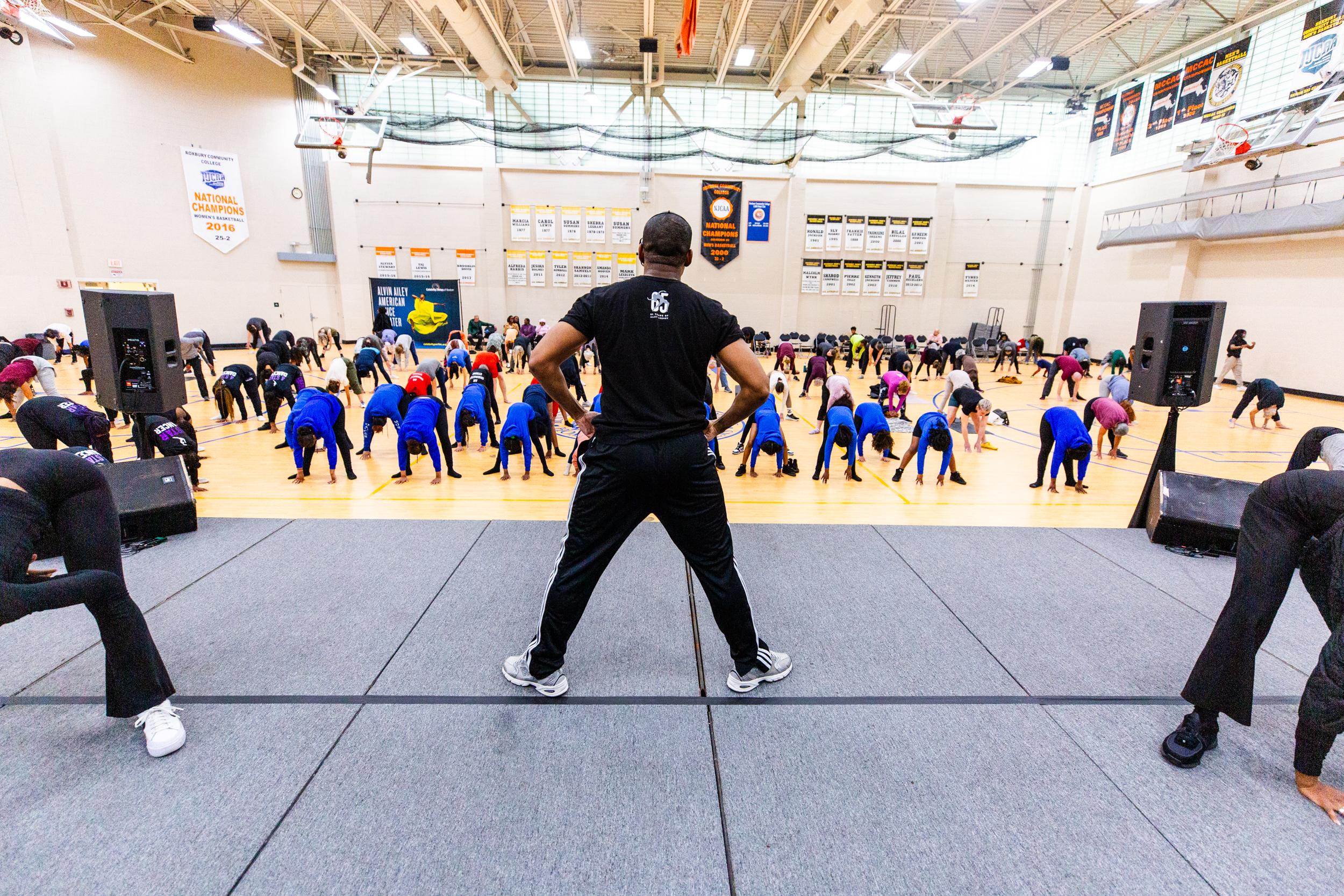  A male instructor in a black t-shirt and track pants stands on a stage with hands on hips, facing a large group of participants stretching in a gymnasium. The participants are wearing various colors, predominantly blue. The gym walls are decorated with banners and signs.