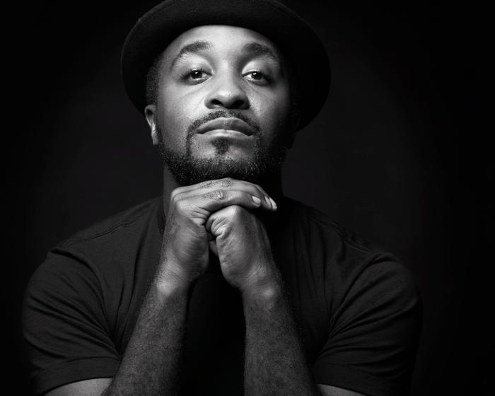 A black and white headshot of a black man. He is wearing a beanie and his head is resting on both of his hands clasped.