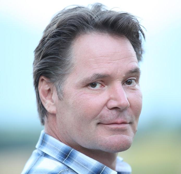 A headshot of a main in a field. He is wearing a blue checkered collared shirt.