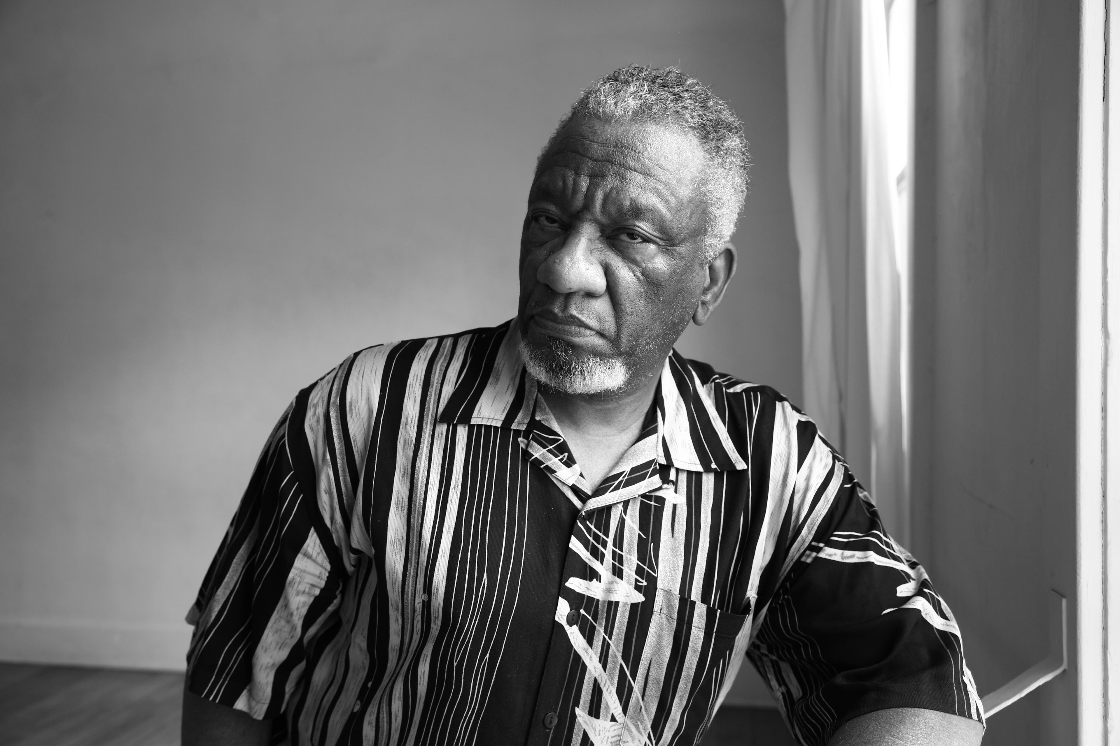 A black man sits with his elbow resting on a table. He's in a striped collared shirt. The photo is in black & white.