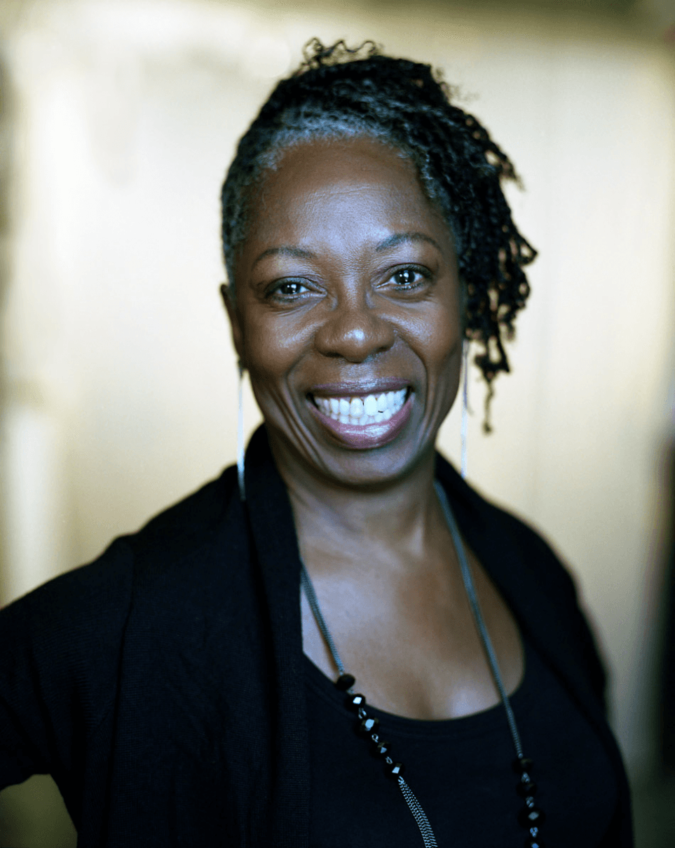 A black woman smiles in a headshot. She is wearing a black top and has glasses around her neck.