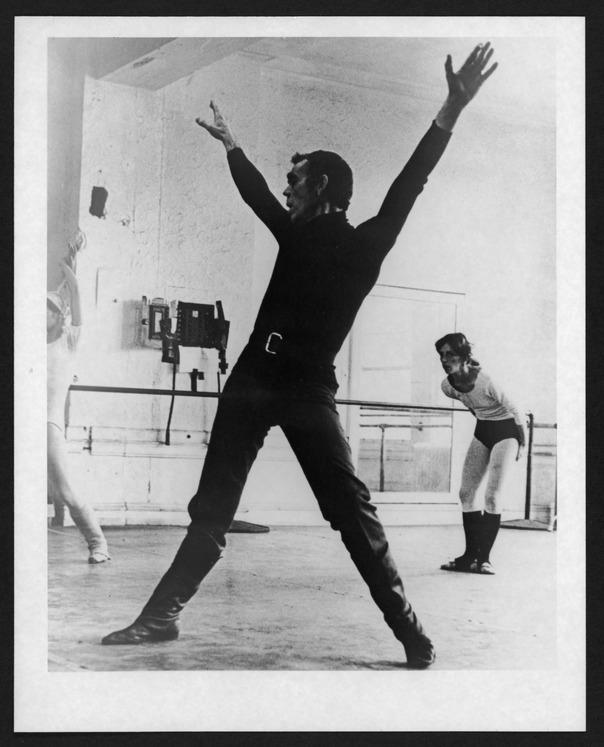 An old photo of a man in a dance room. His arms are in a V and his legs are apart.