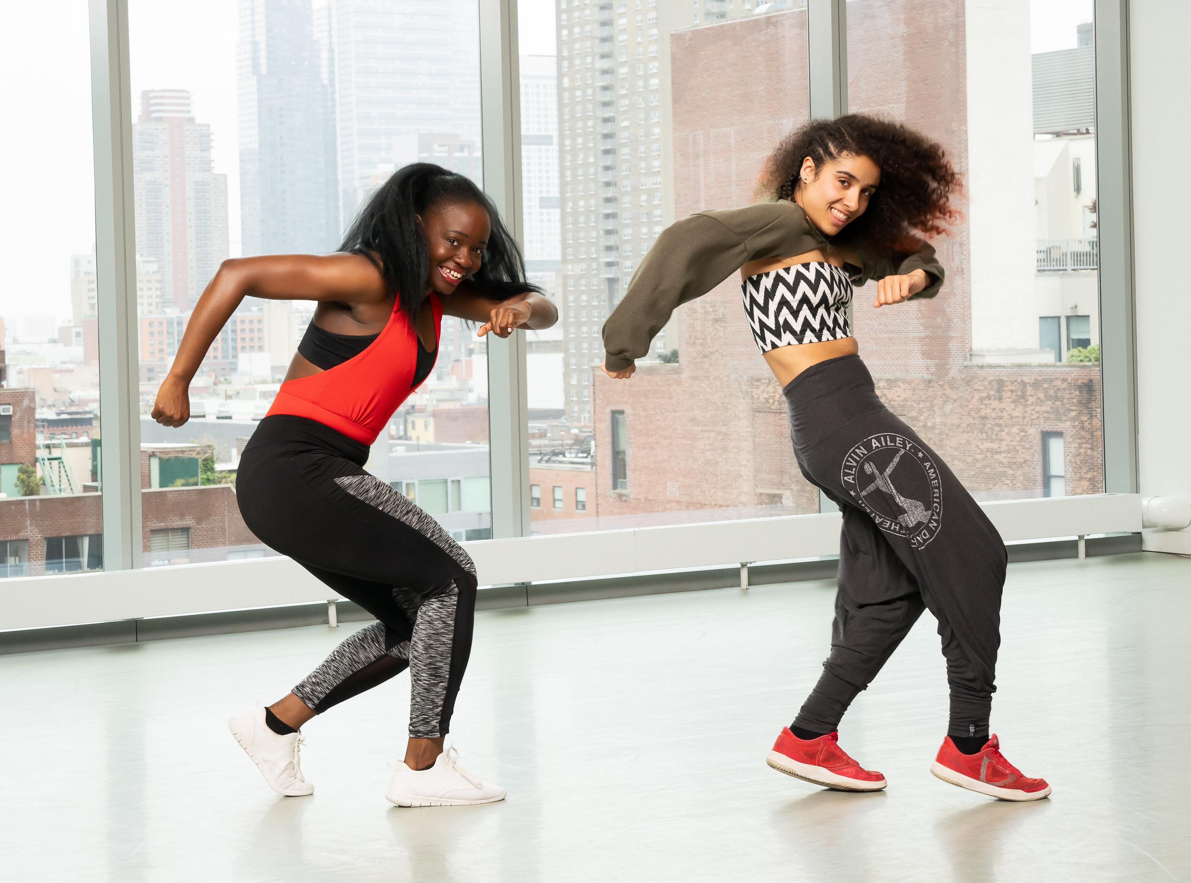 The dancers are engaged in dynamic and expressive movements, showcasing the vibrant energy of AfroDance. The studio's large windows provide a backdrop of the cityscape, adding a modern urban feel to the scene. The dancers are dressed in comfortable athletic wear, suitable for their energetic routine. 