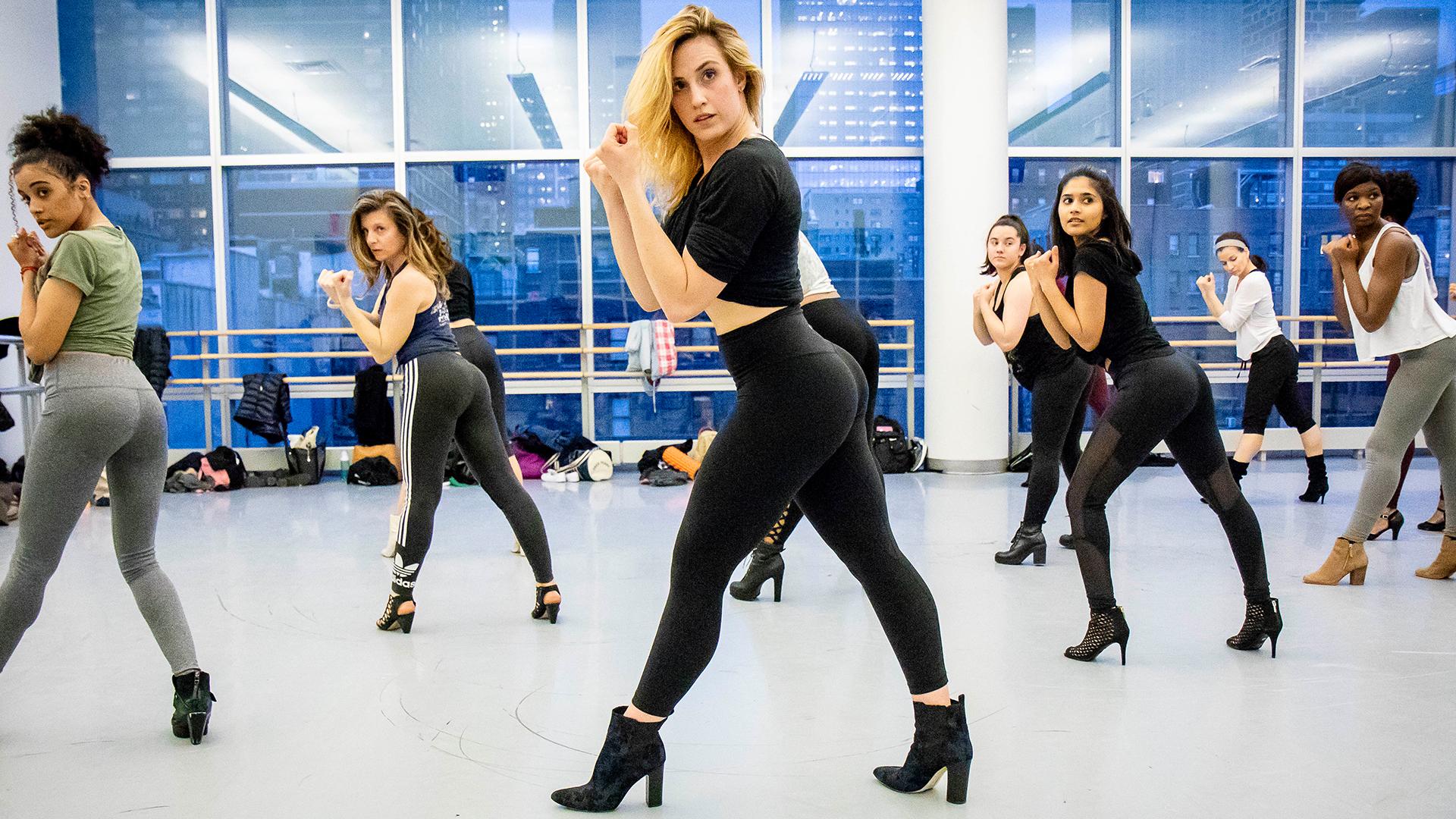 The dancers, dressed in stylish workout attire and high-heeled shoes, are captured in a powerful and confident pose, highlighting their strength and grace. The studio's large windows reveal a cityscape backdrop, adding an urban vibe to the energetic atmosphere. The instructor leads the class with precision, encouraging the dancers to embody the fierce and empowered style characteristic of Heels dance. 