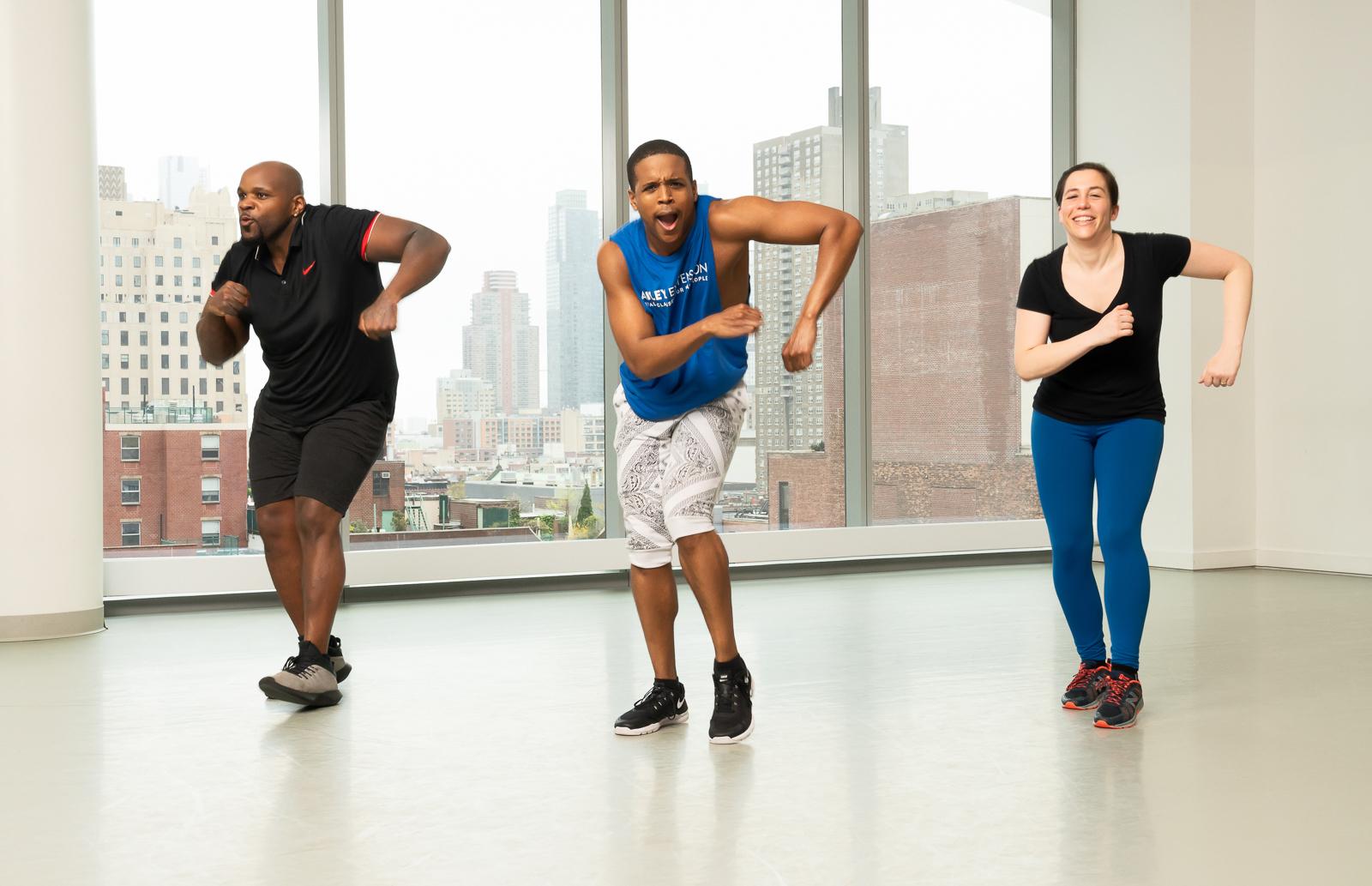 The participants, dressed in athletic wear, display dynamic and synchronized movements, emphasizing the high-energy and fun atmosphere of the class. The backdrop of large windows showcases an urban cityscape, adding to the lively ambiance. This class focuses on combining cardio exercises with hip hop dance moves, providing a full-body workout that's both engaging and effective. 