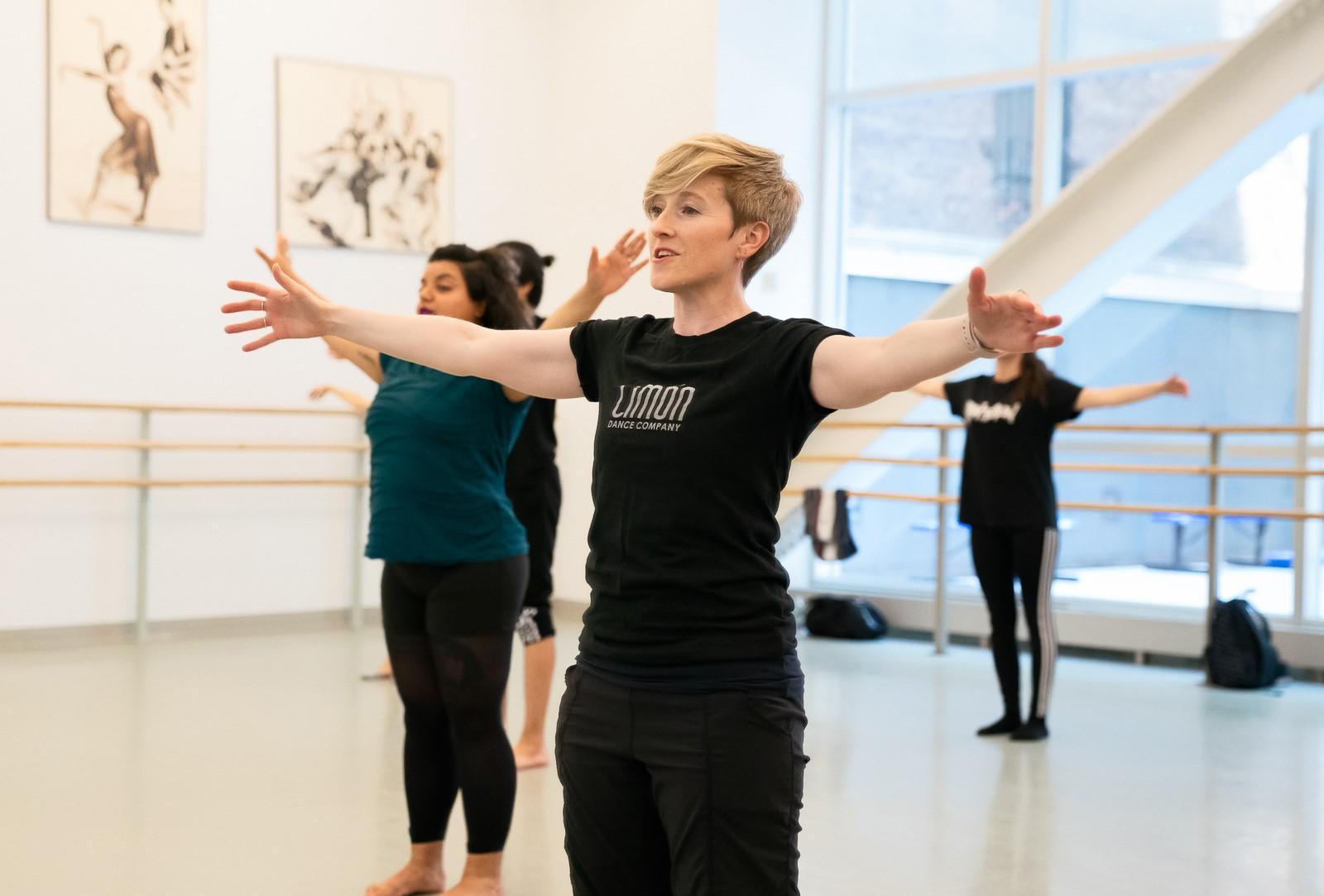 The instructor, wearing a black shirt with "Limon Dance Company" printed on it, is leading the class with her arms extended. The participants in the background follow along, also extending their arms, and appear focused and engaged in the movement. 