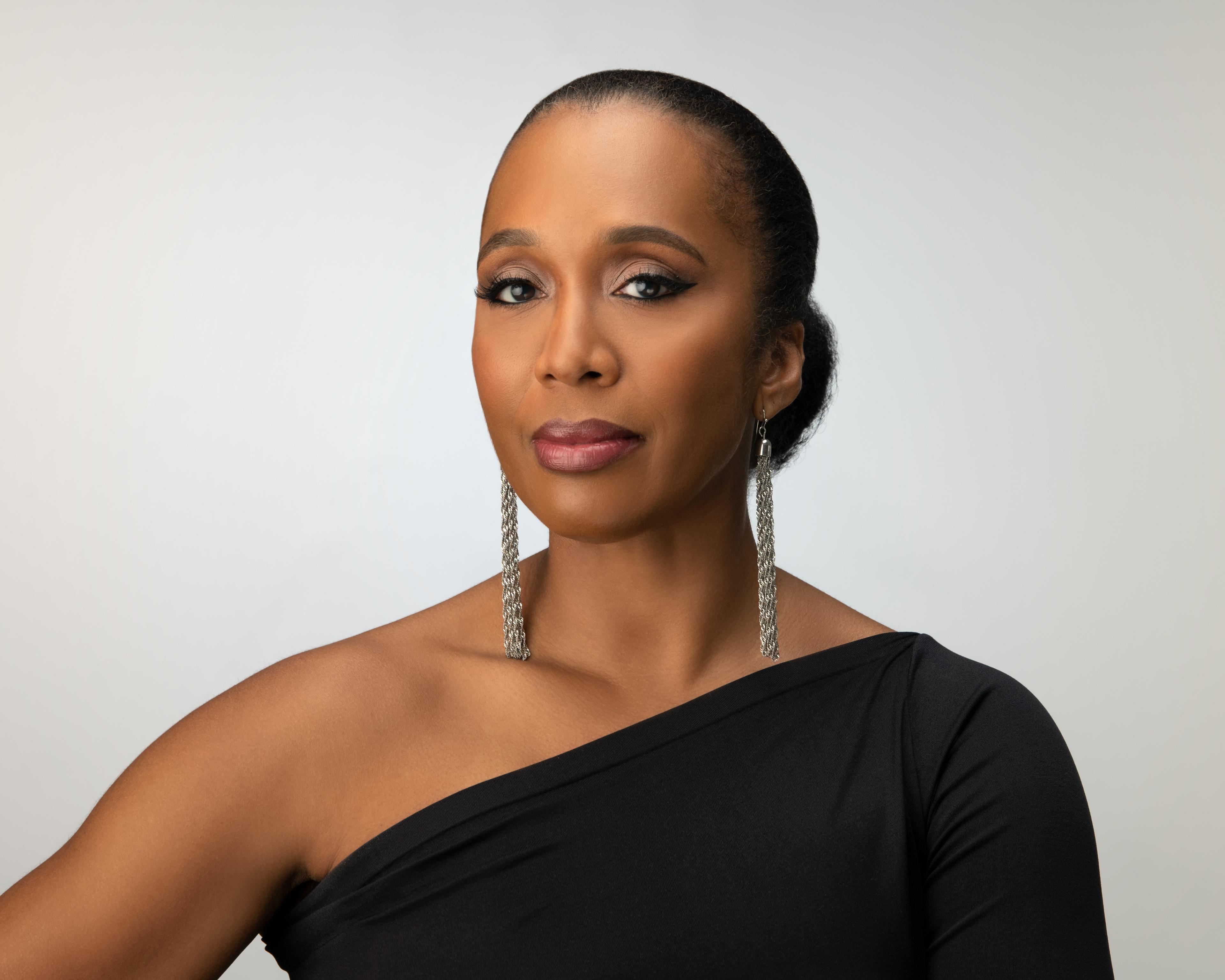 Portrait of Francesca Harper, Artistic Director of Ailey II. She is wearing a black, one-shoulder top and long, silver earrings. Her hair is pulled back, and she has a calm expression. 