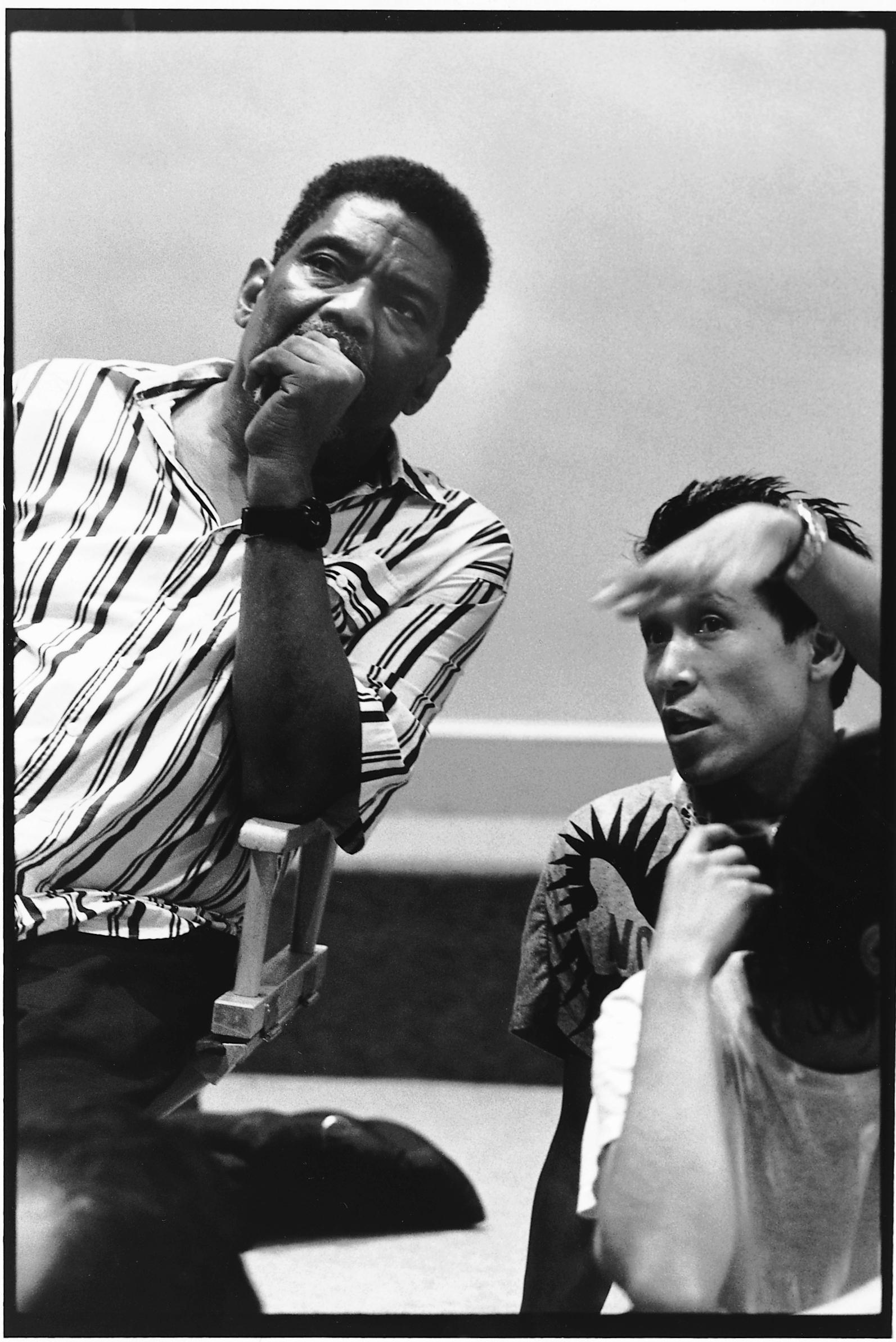 Black and white photo of Alvin Ailey and Masazumi Chaya in rehearsal, early 1980s. Ailey, in a striped shirt, is seated with his hand on his chin, looking thoughtful. Chaya, to the right, gestures as he speaks. 