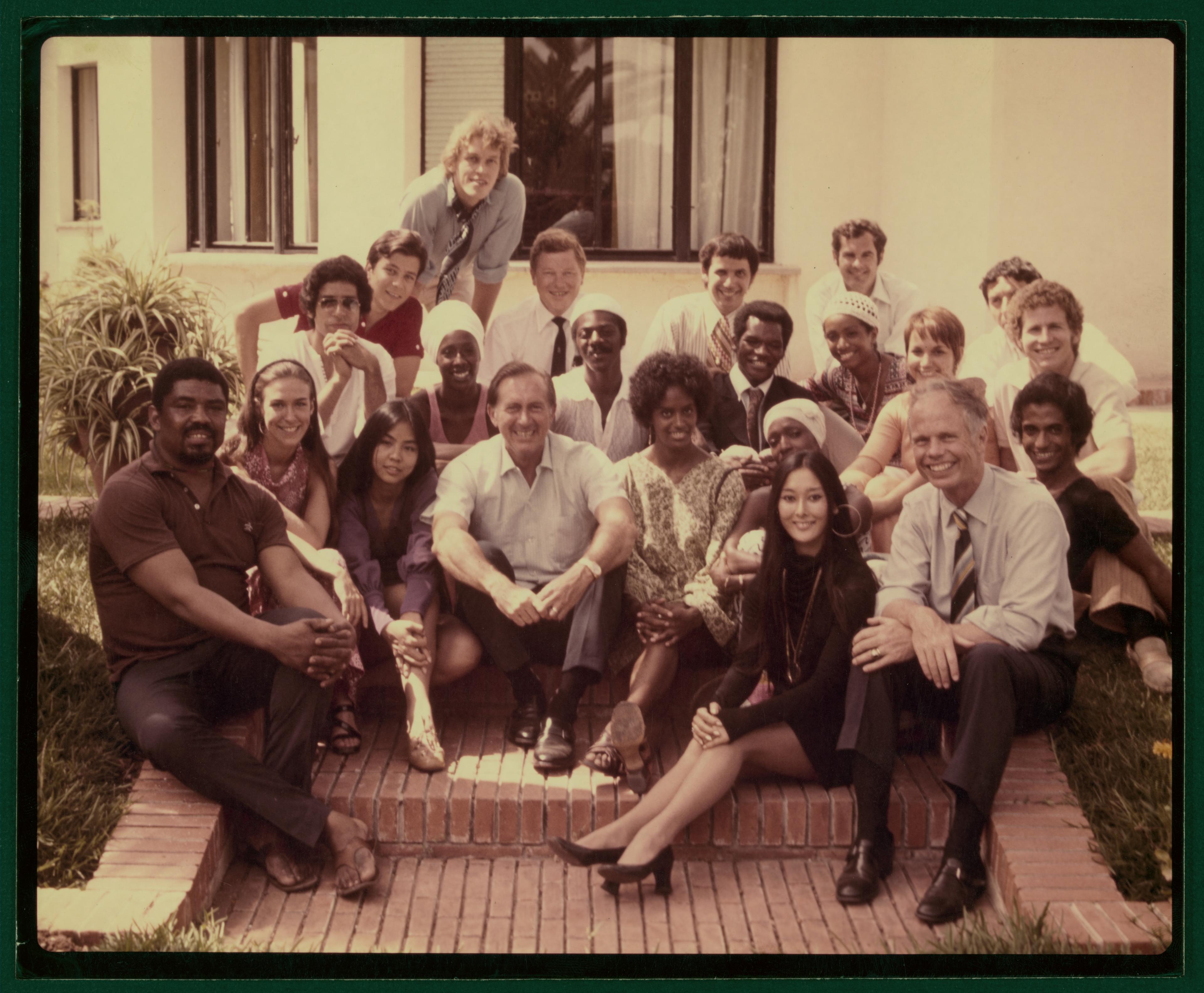 Group photo of Alvin Ailey, Judith Jamison, Sylvia Waters, and company members at Casa Blanca, American Embassy, during the North African Tour, 1970. The group is seated on steps and arranged in a relaxed manner, smiling at the camera. They are outdoors, in front of a building with large windows. 