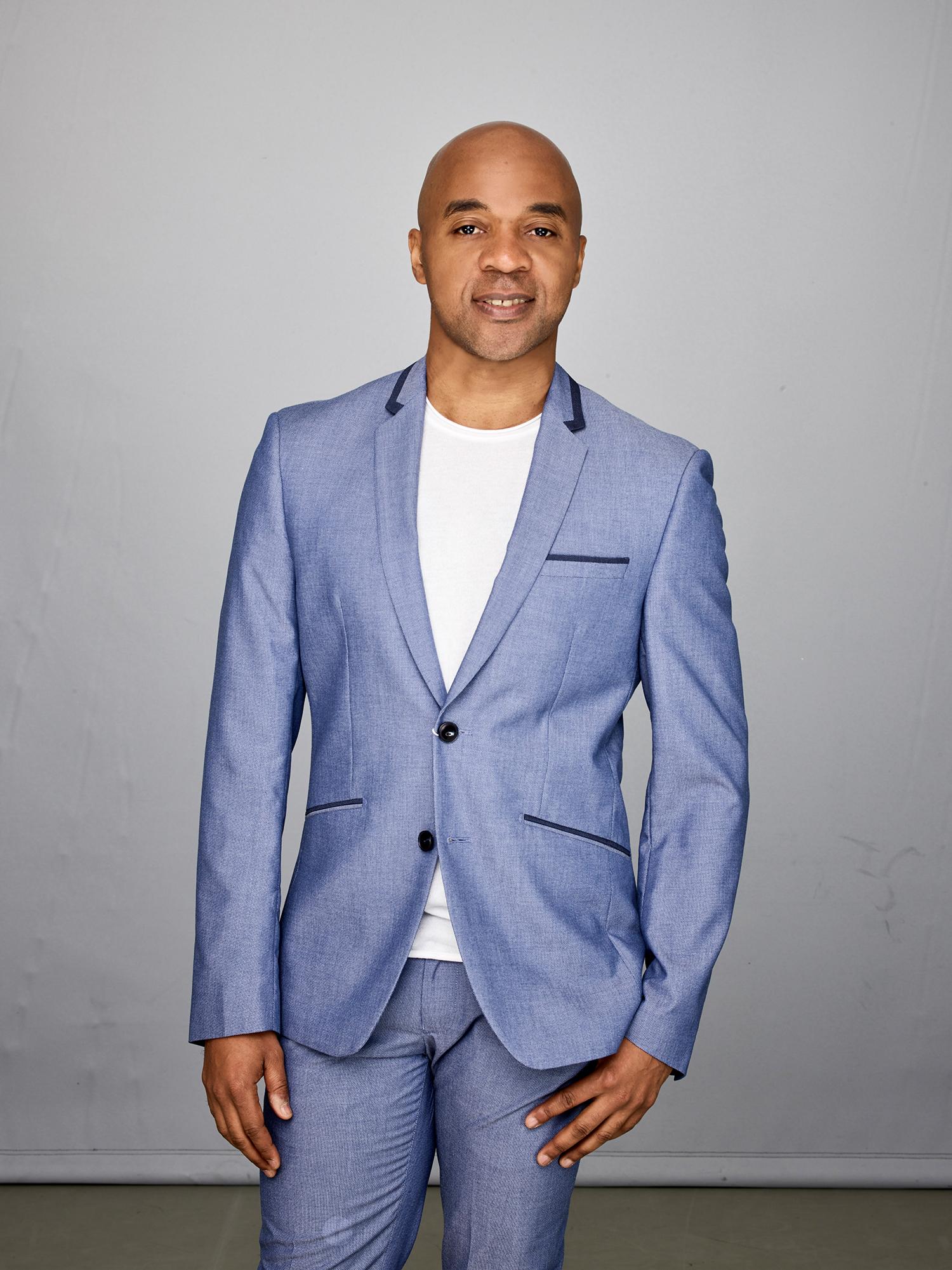 Portrait of Matthew Rushing, Associate Artistic Director. He is wearing a light blue suit with a white shirt, standing against a plain background, smiling slightly at the camera. 