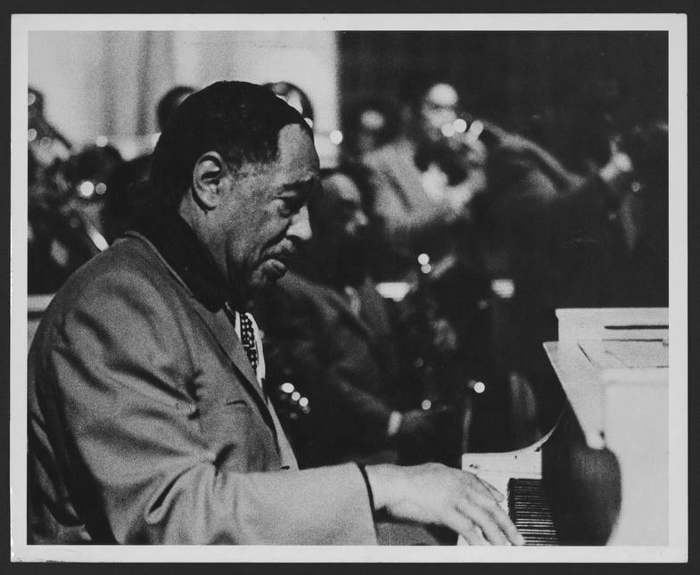 Black and white photo of Duke Ellington playing the piano. He is focused on the keys, dressed in a suit. In the background, a band is visible, with musicians playing various instruments. Photo by Stanley Dance, 1963.