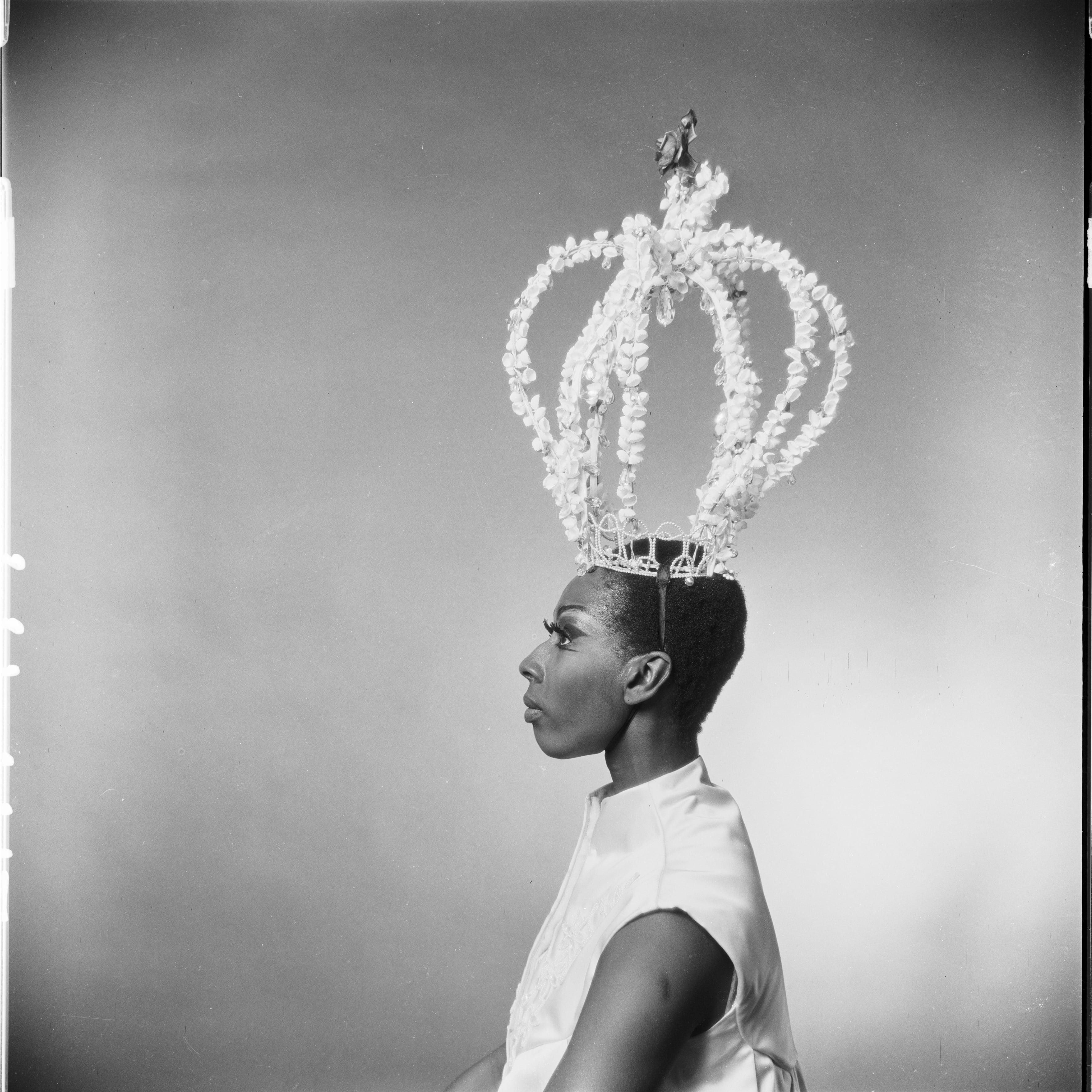 Black and white profile photo of Judith Jamison wearing a large, ornate crown. She is dressed in a sleeveless top and gazes forward with a calm expression. The background is plain, emphasizing her regal appearance. The photo is from 1967.