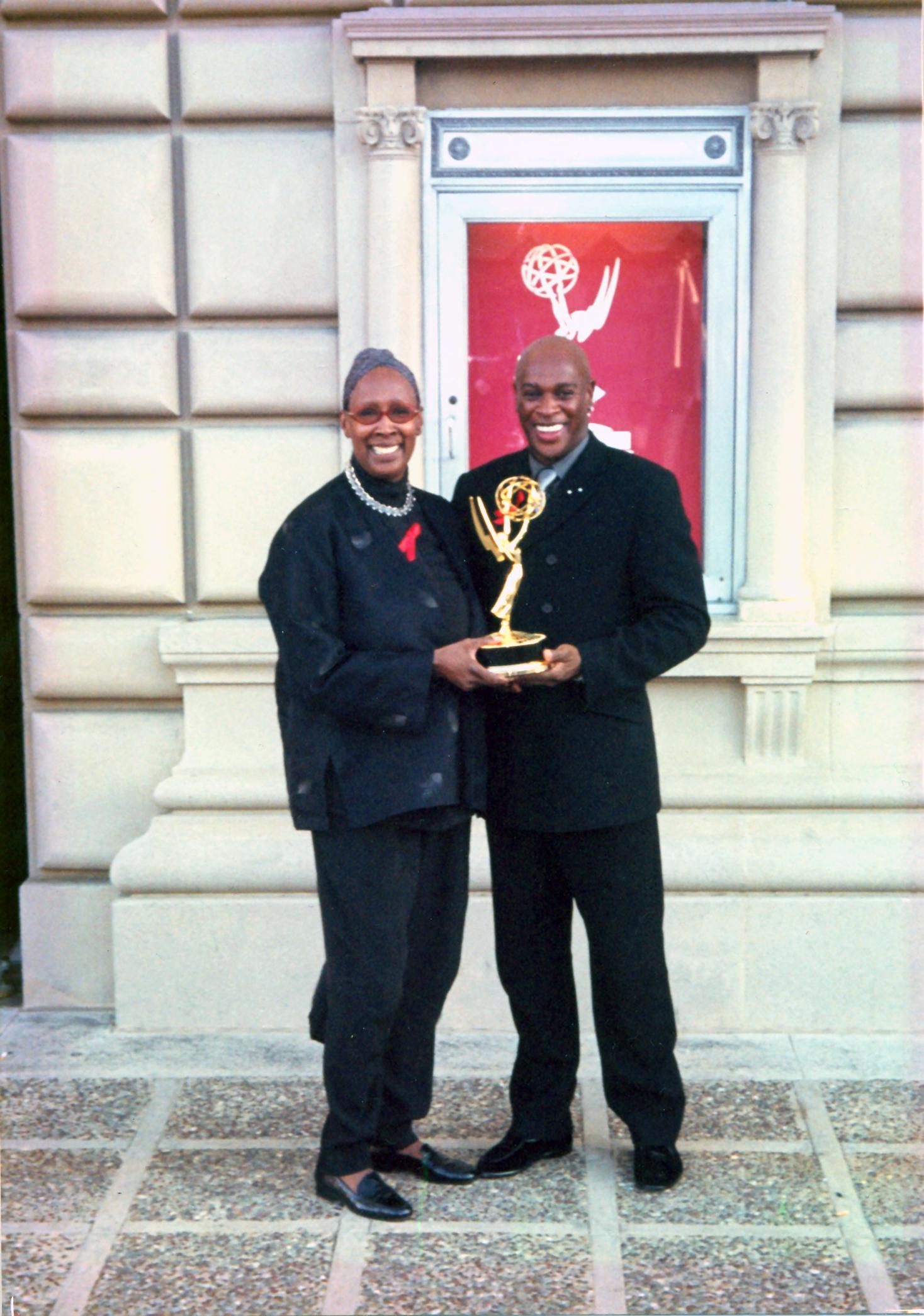  Judith Jamison and a colleague holding an Emmy award outside a building. Both are smiling and dressed in dark attire. The background features a red poster with the Emmy logo. The photo is from 1999.